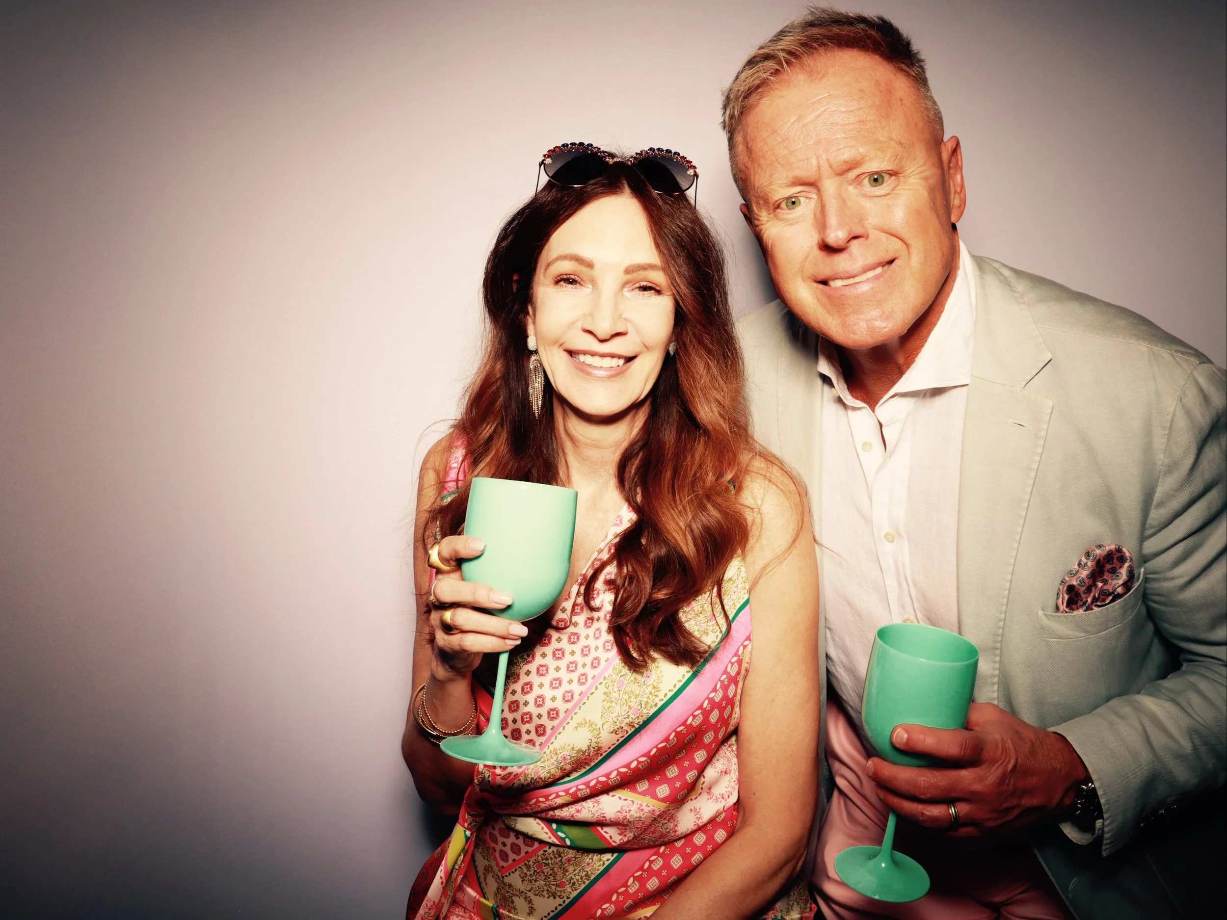 A smiling woman with long red hair wearing a colorful top and sunglasses on her head, holding a green wine glass. A smiling man with short blonde hair wearing a light blazer, white shirt, and pink pants, also holding a green wine glass, standing besi
