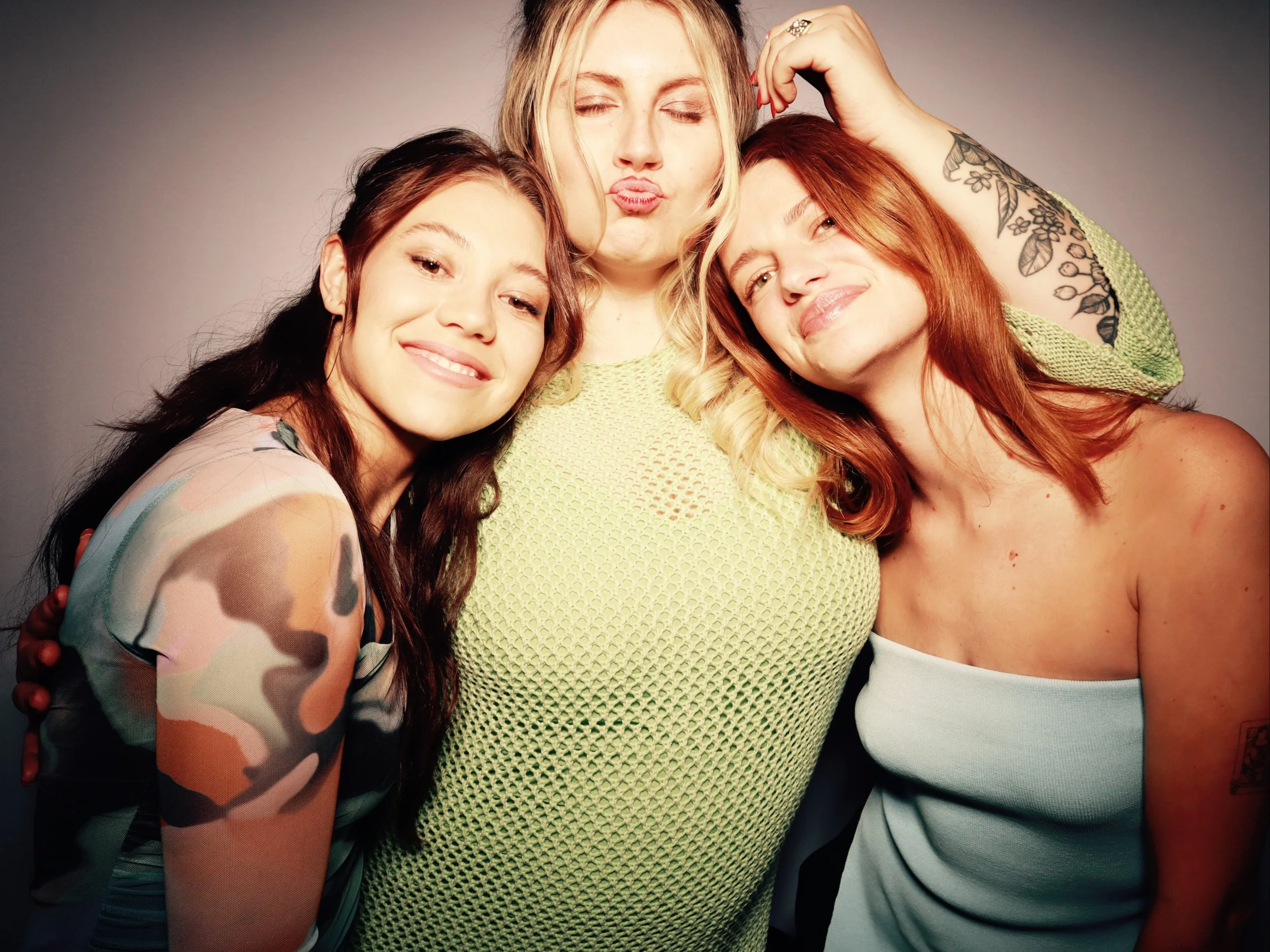 Three women hugging and smiling at the camera, with one woman making a kissy face, against a plain gray background.