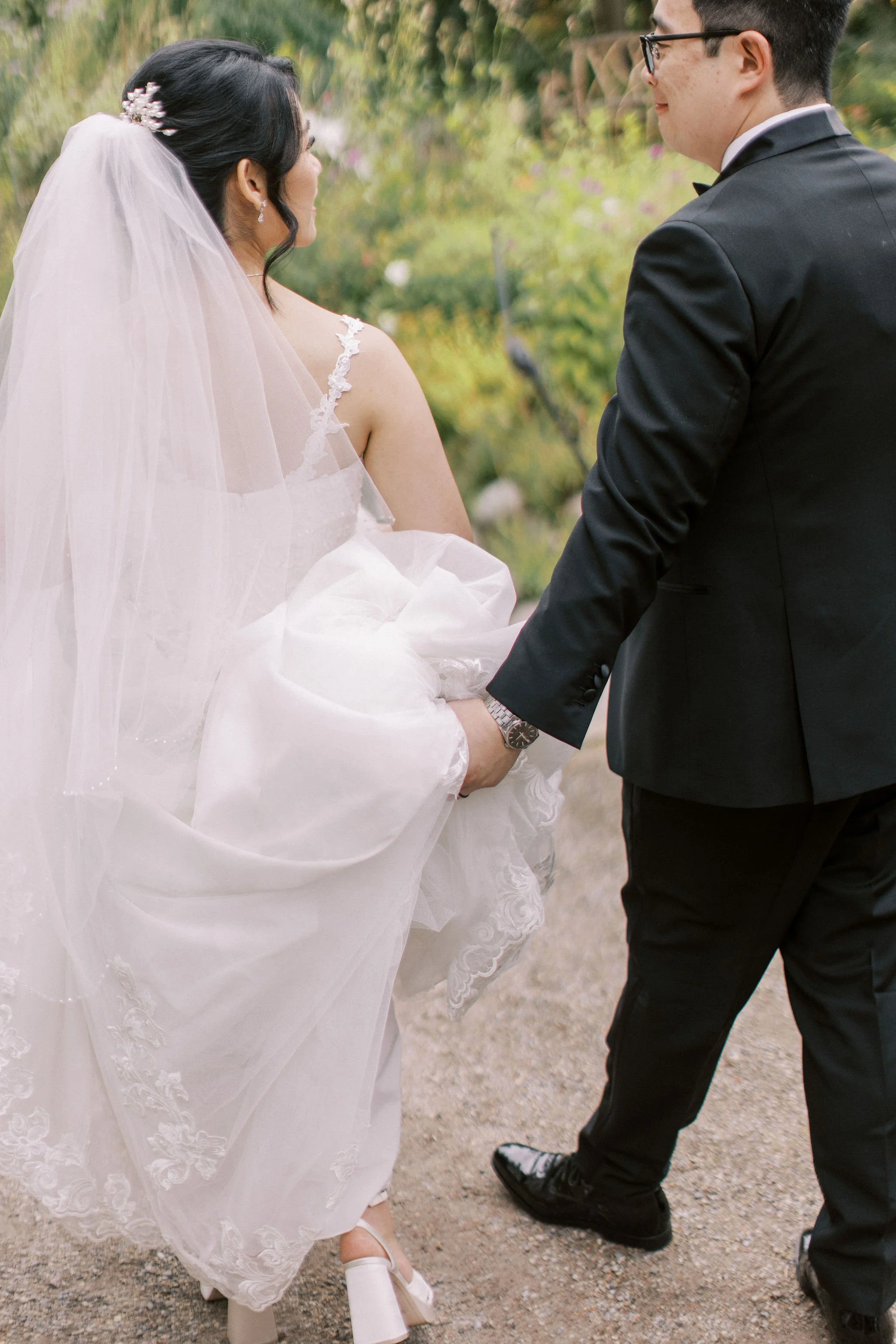 A bride and groom holding hands outdoors during their wedding, with the bride in a white dress and veil, and the groom in a black suit and glasses.