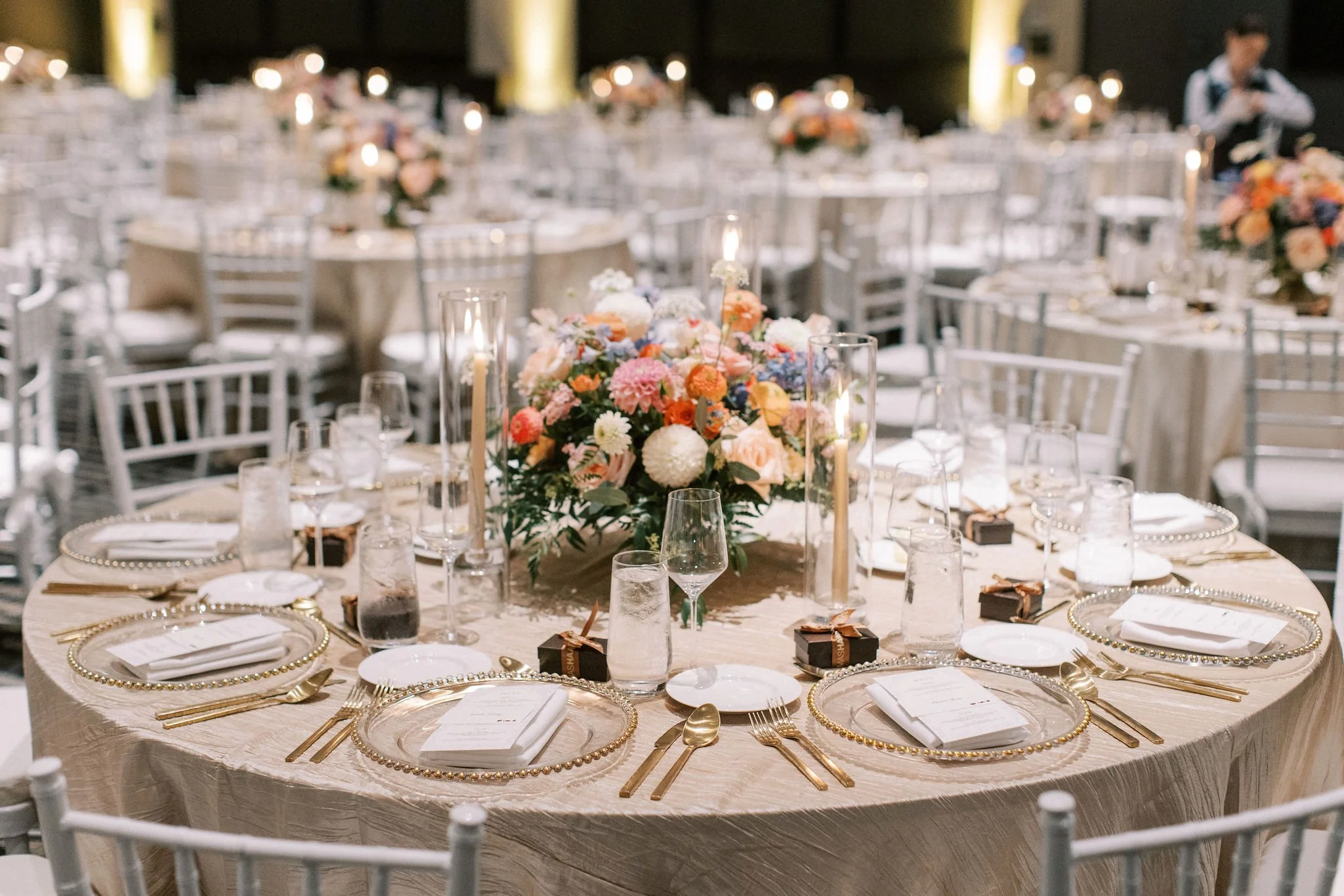 Round event table decorated with a large floral centerpiece, tall candle holders, and place settings with gold flatware, white napkins, and clear glassware, in a banquet hall.