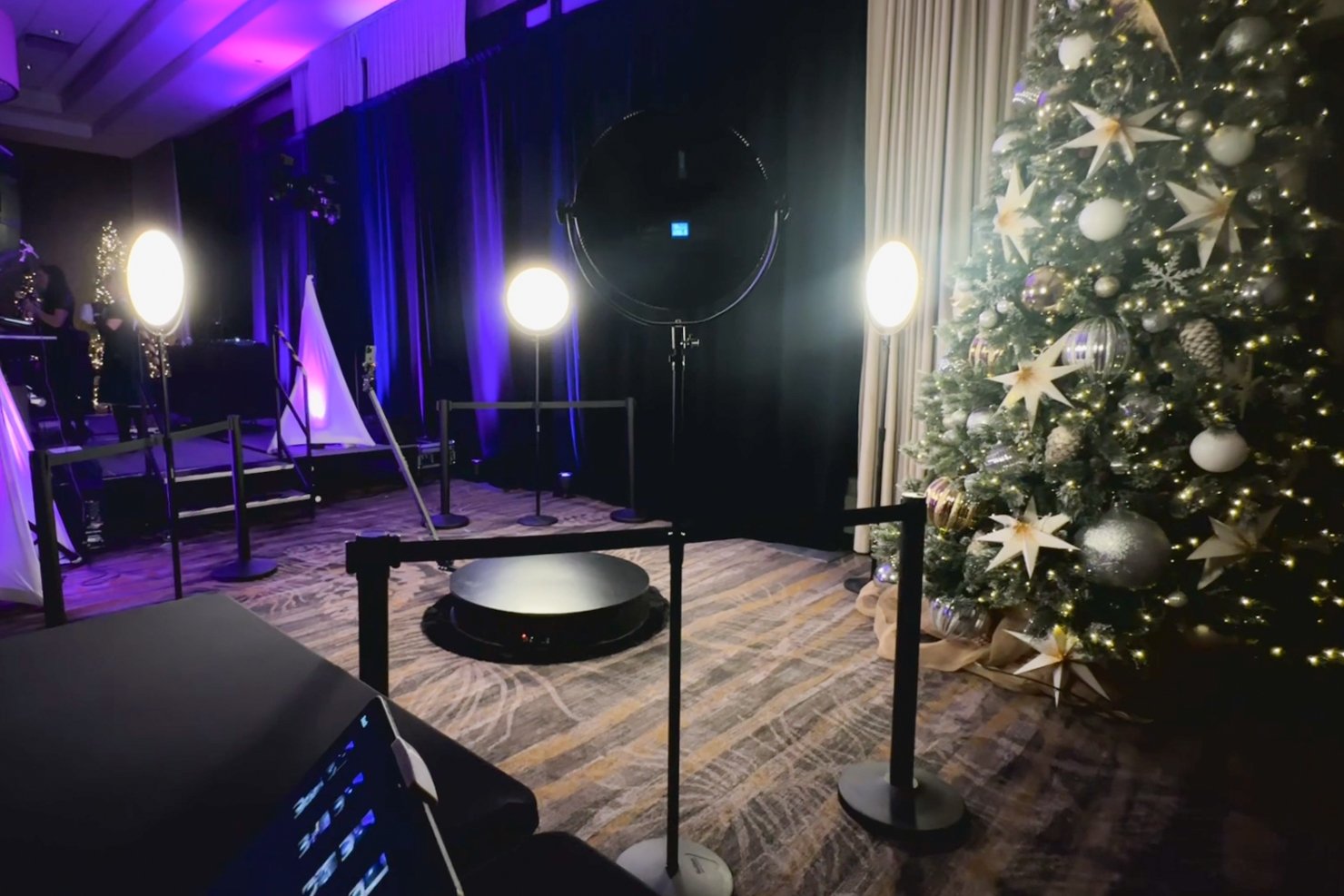 Holiday photo setup with Christmas tree decorated with silver and gold ornaments, white stars, and lights on the right side. Stage with bright lights and purple curtains in the background, and some people preparing in the dark.