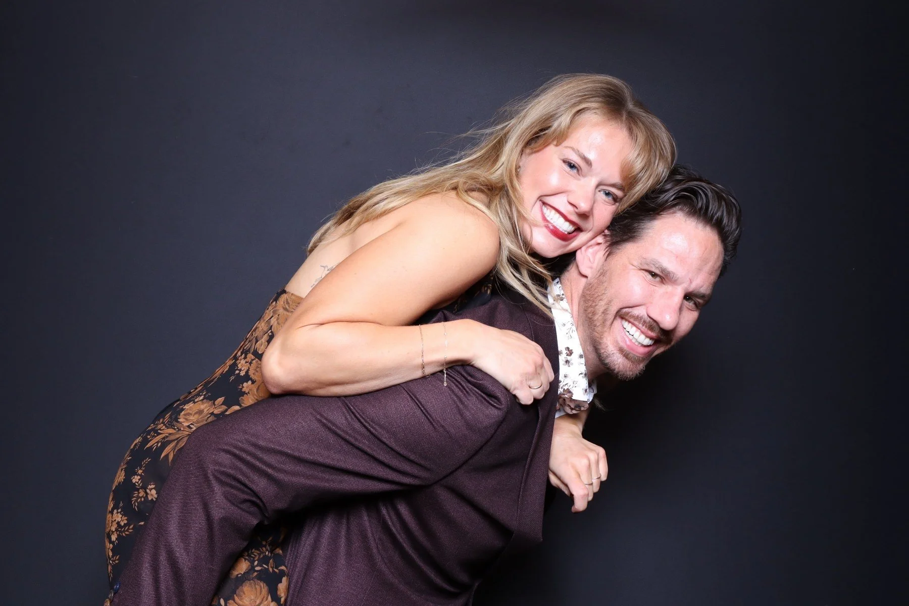 A couple smiling, the woman on the man's back, against a dark background.