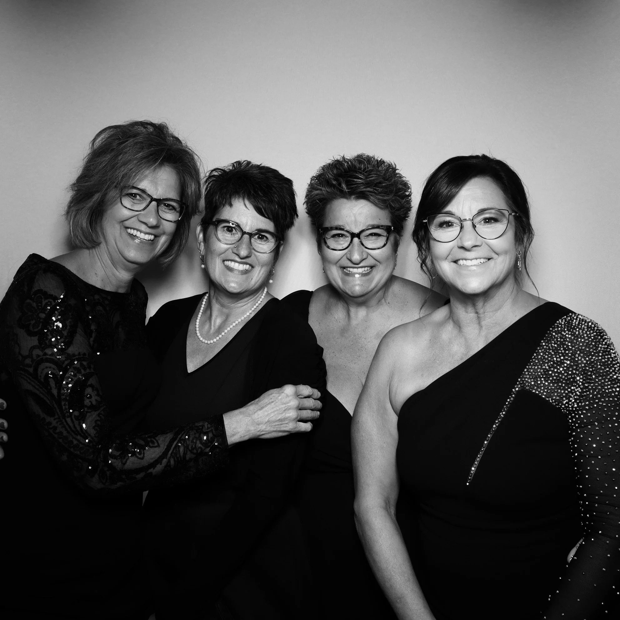 Black and white photo of four women wearing elegant dresses and glasses, smiling in front of a plain background.