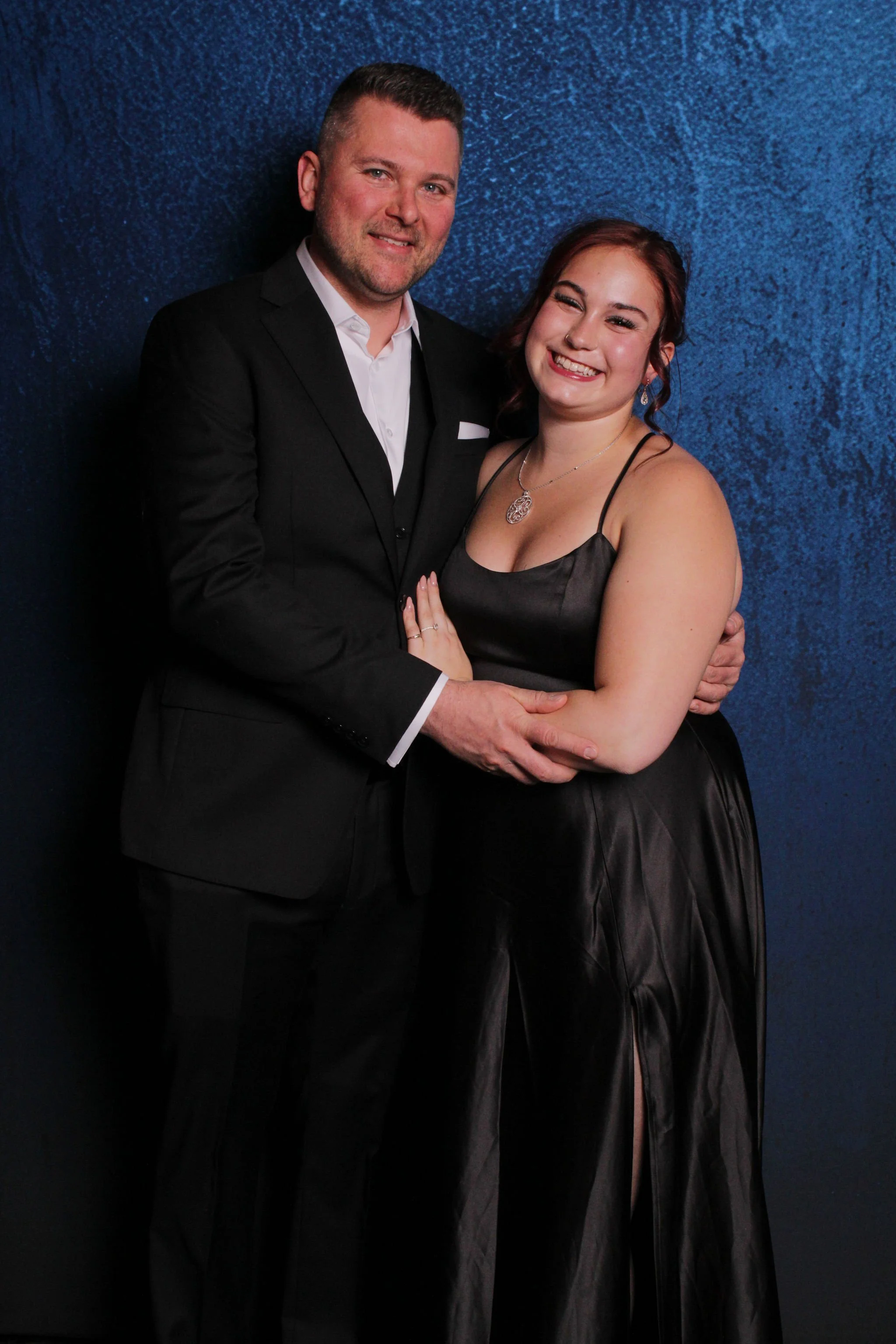 A man and woman dressed formally, smiling and embracing, standing against a dark blue textured backdrop.