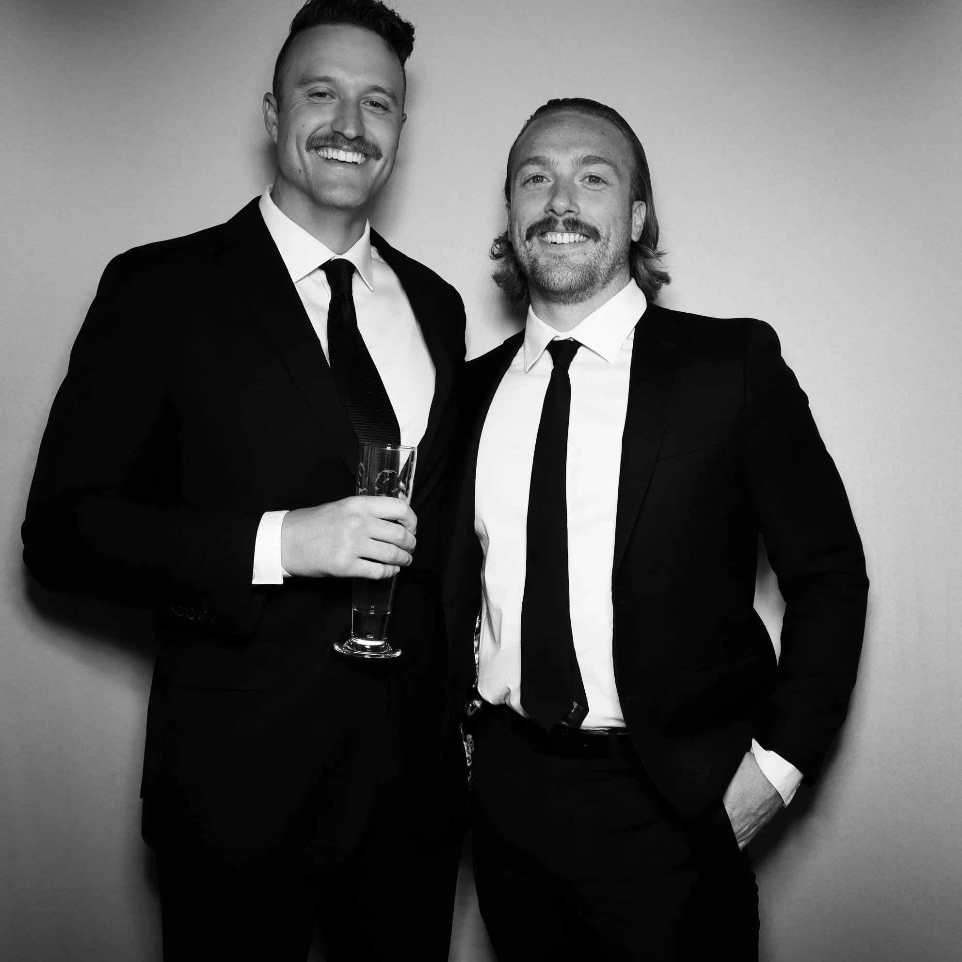 Two men in suits smiling at the camera, one holding a glass of champagne, black and white photo.