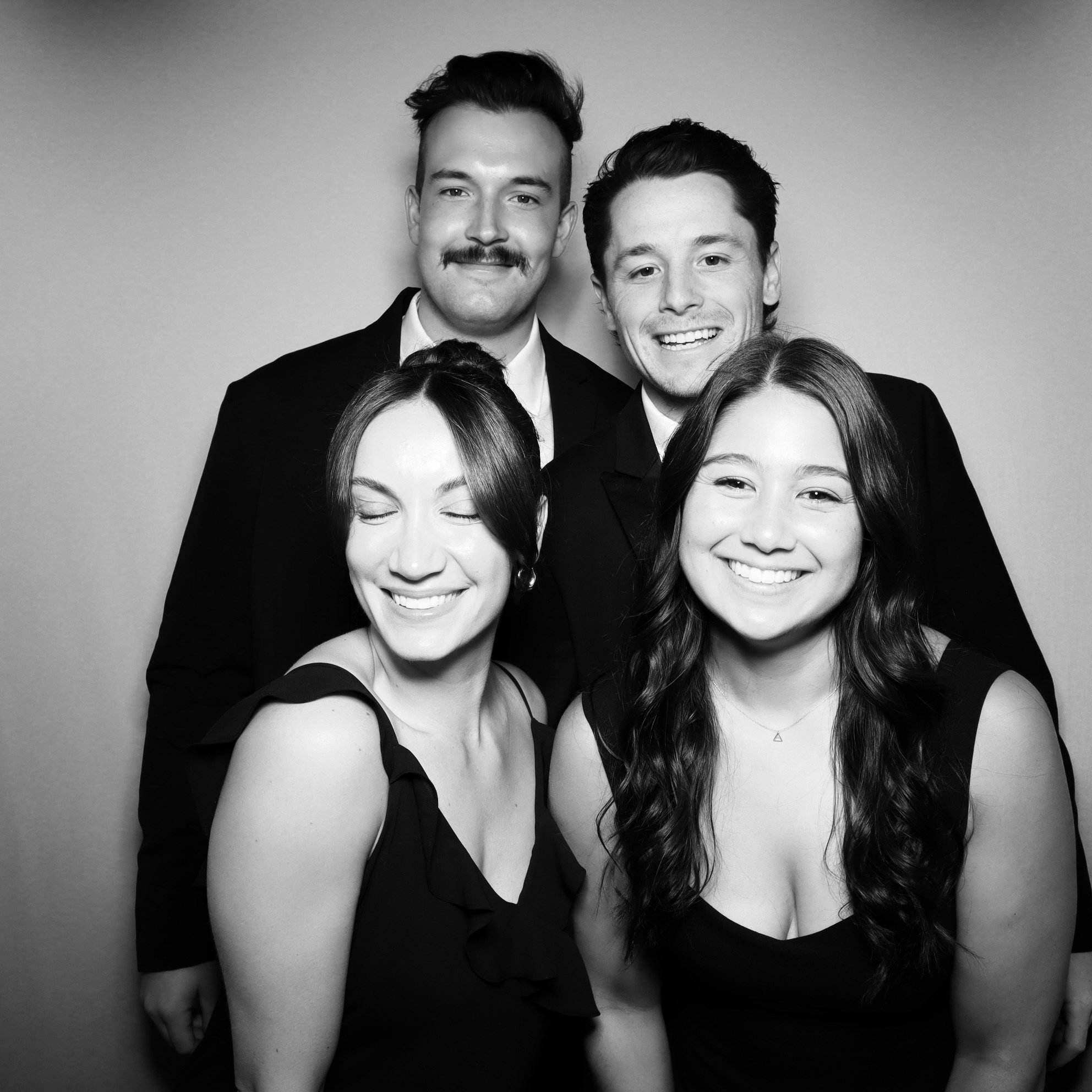 A black and white photo of five young adults smiling and posing together against a plain background, dressed in formal attire.