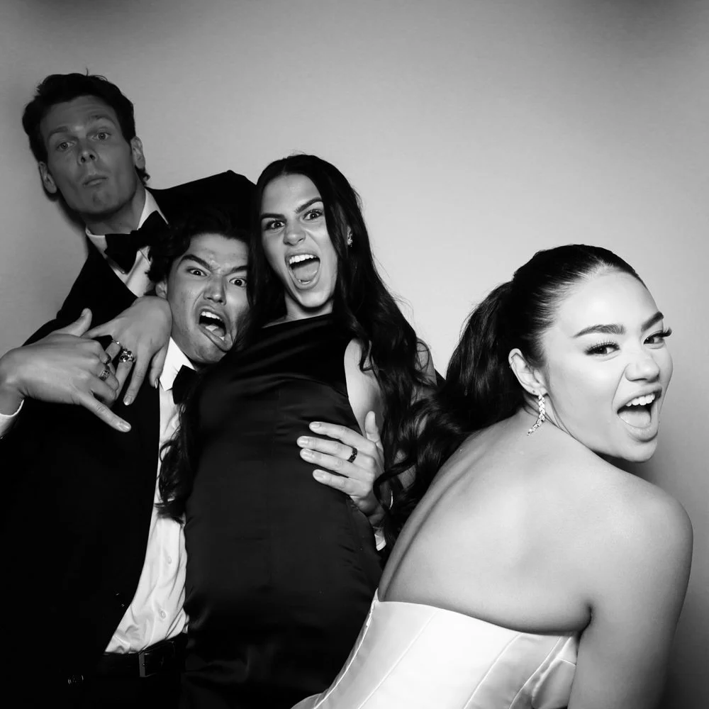 Black and white photo of four young adults, three men and one woman, making playful and energetic expressions at a party or event. The woman is in a strapless dress, and the men are in formalwear.