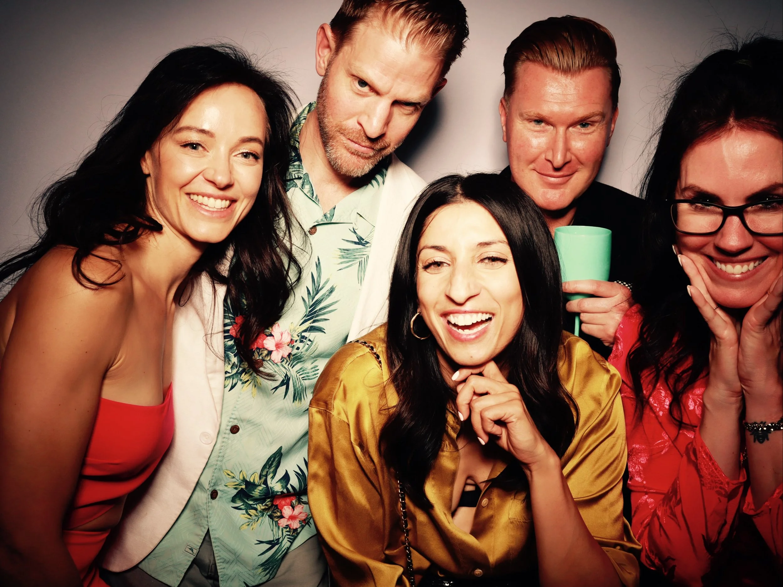 Group of five friends smiling and posing together at a party or gathering, with one holding a teal mug.