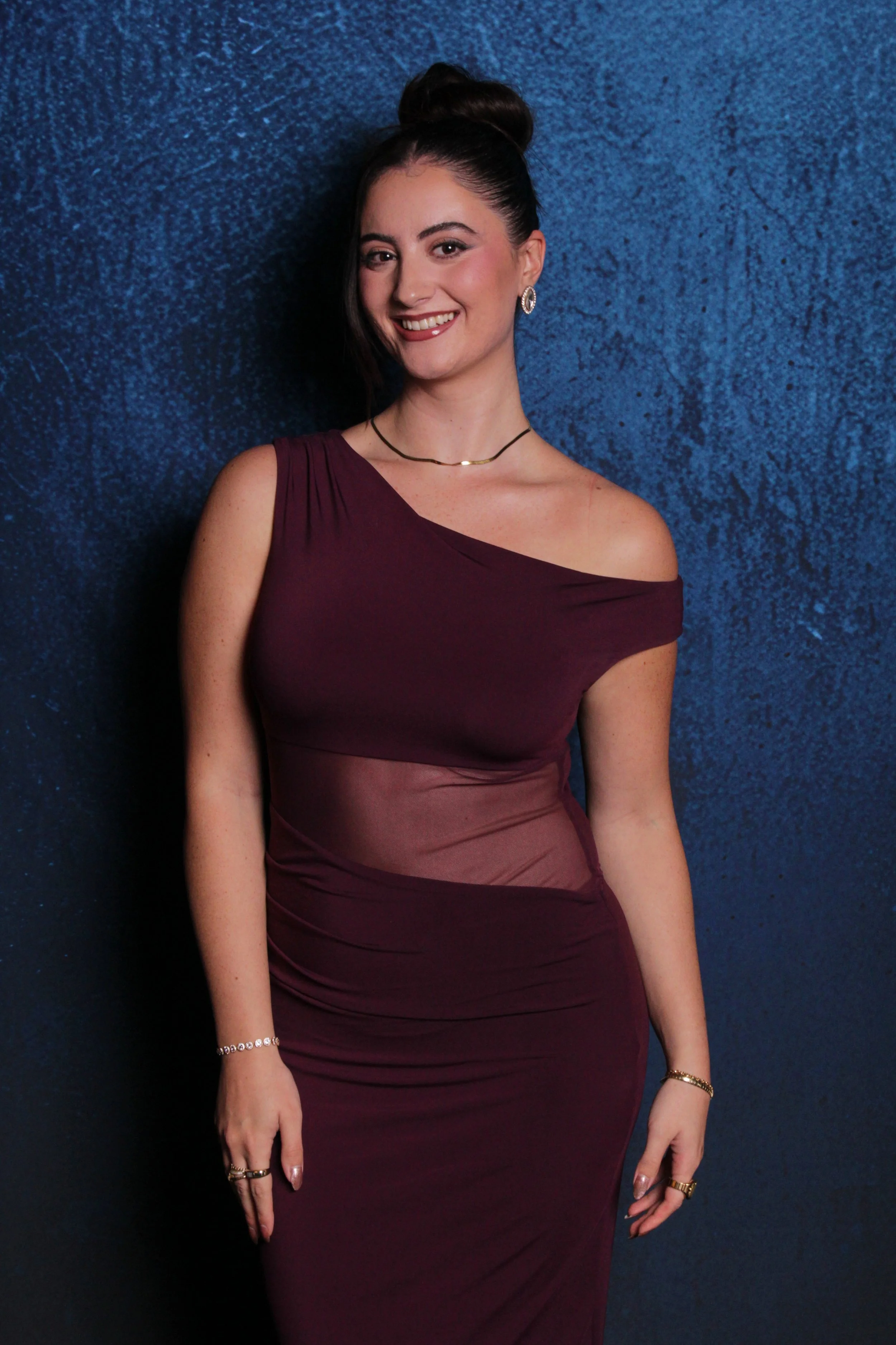 A woman with long dark hair styled in a high bun, smiling, standing against a dark blue textured wall, wearing a one-shoulder plum-colored dress with a sheer panel, accessorized with jewelry.