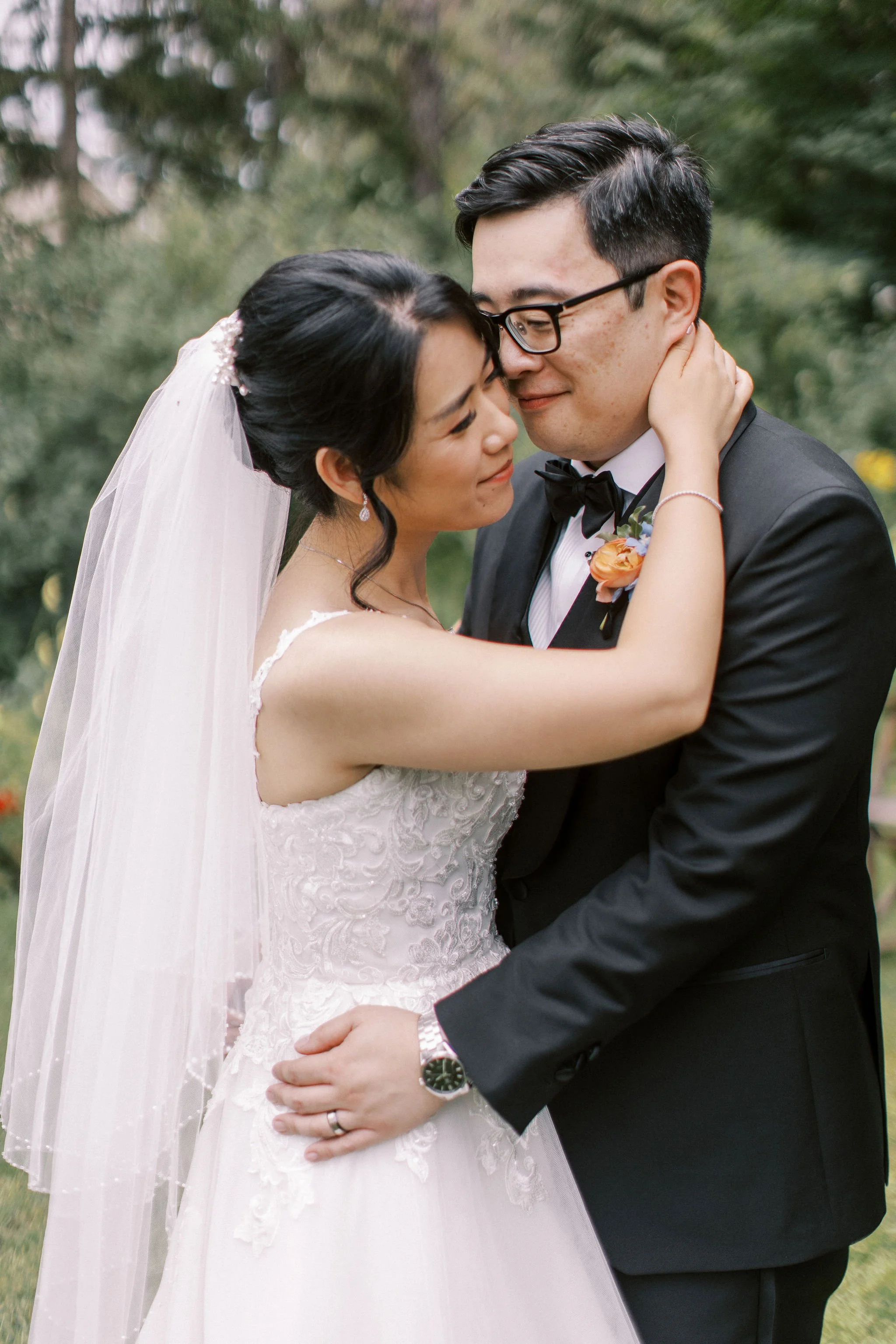 A bride and groom embrace outdoors, with the bride wearing a white lace wedding dress and veil, and the groom in a black tuxedo with glasses, both smiling gently.