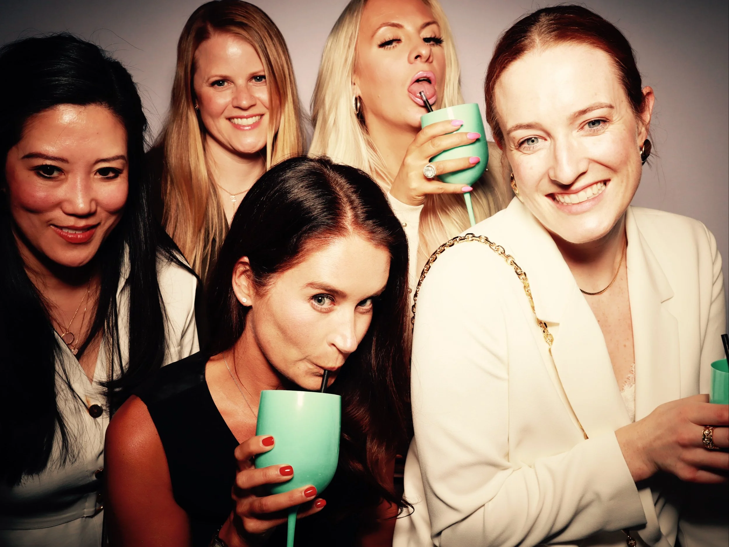 Group of six women at a social gathering, some sipping drinks, smiling, and posing for the camera, with neutral background.