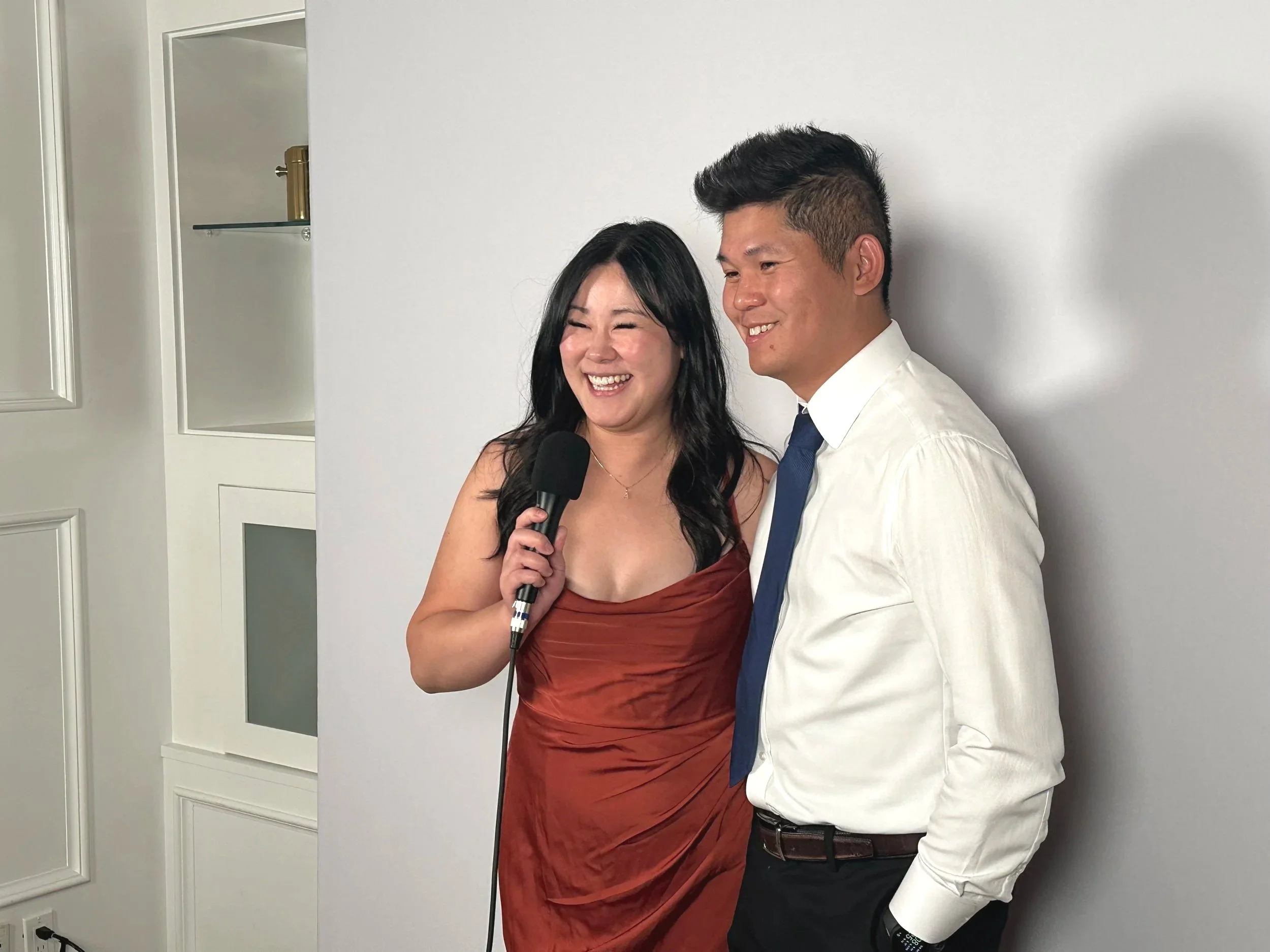 Two people standing close together, one woman holding a microphone, smiling, and another man smiling, both dressed in formal attire, against a white wall background.