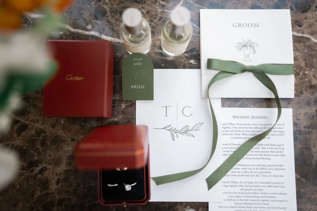 Wedding preparation items arranged on a marble surface, including a Cartier jewelry box with rings, a red box, a green tag labeled 'bride', a white wedding blessing card, a groom booklet with a green ribbon, and bottles of perfume or sanitizer.