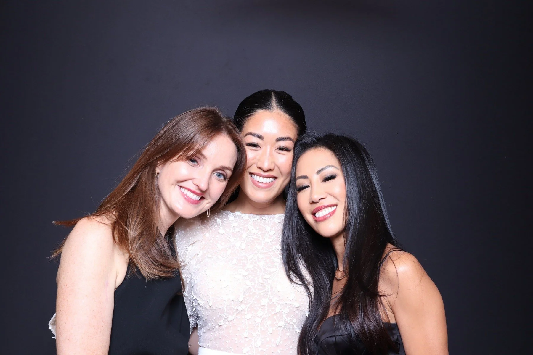 Three women smiling and posing closely together against a dark background.