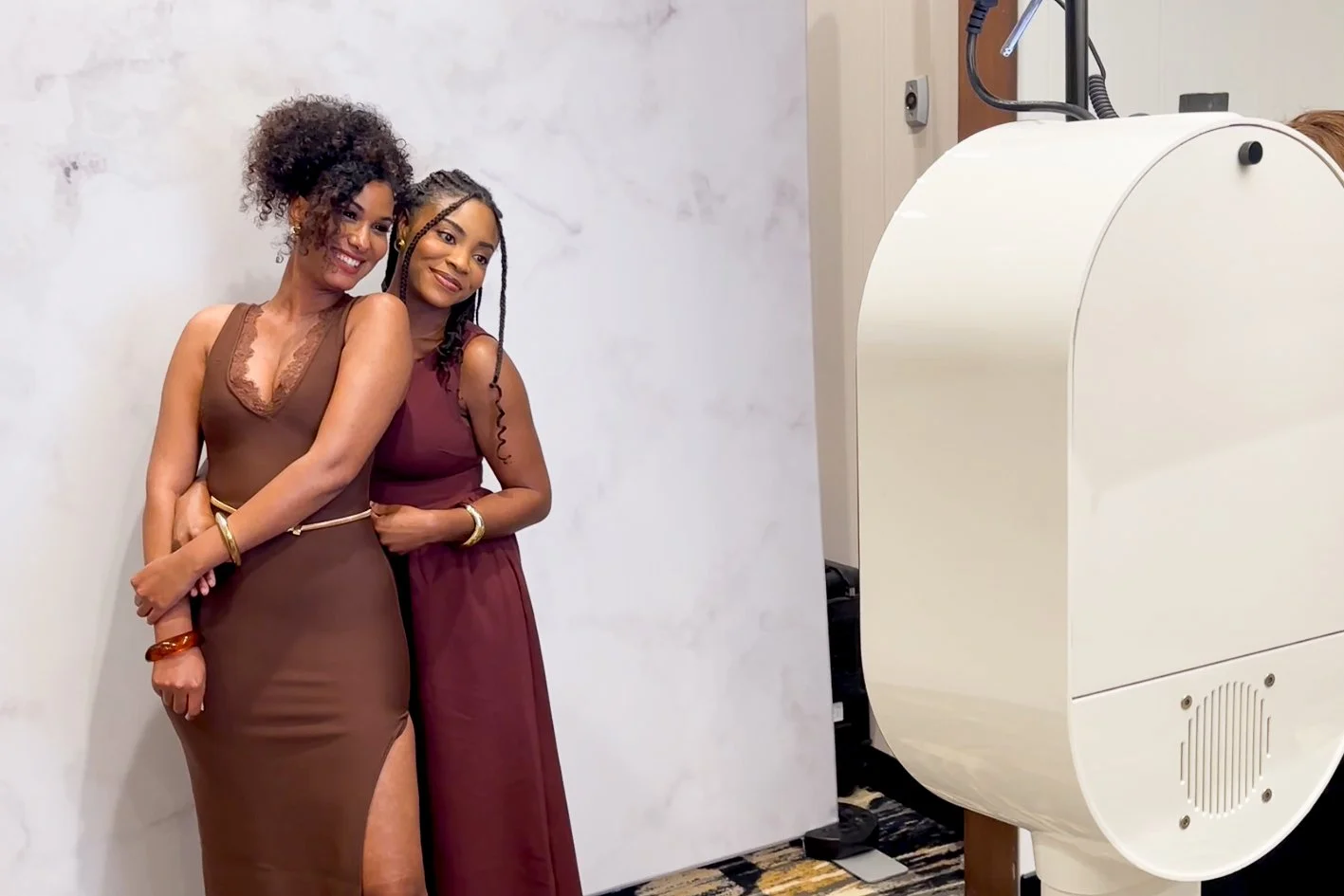 Two women dressed in brown and maroon dresses posing together for a photo, standing next to a large piece of medical imaging equipment in a medical office or clinic.