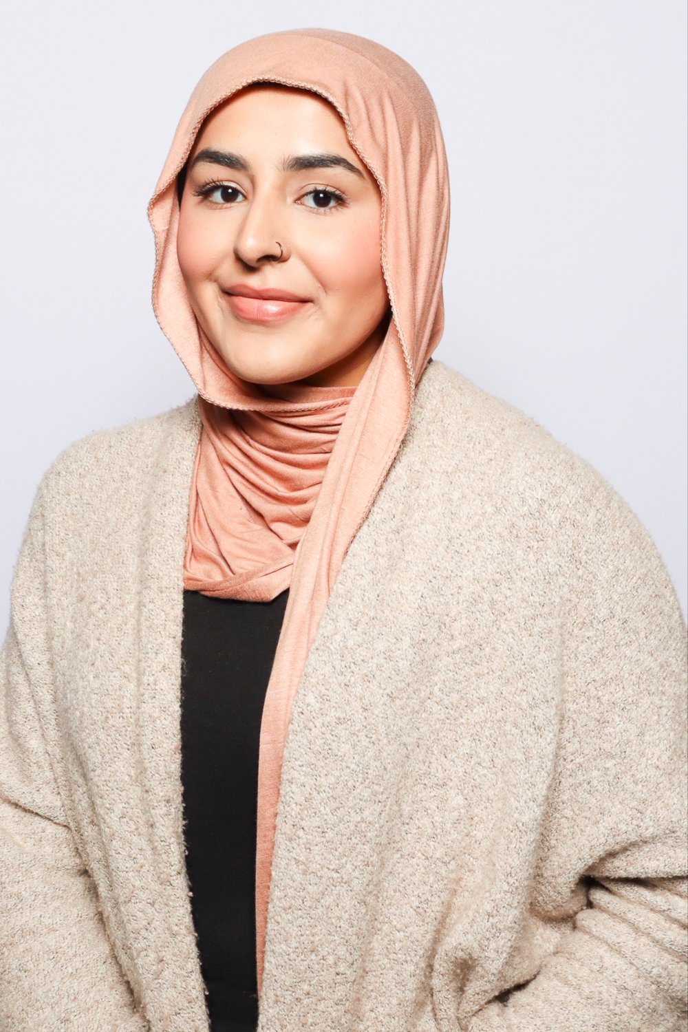 A woman with a light pink headscarf, wearing a beige coat and black top, smiling softly against a plain light background.