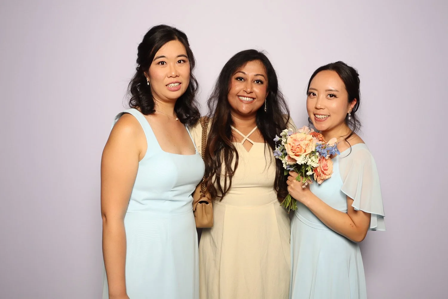 Three women standing together, smiling, with the woman on the right holding a bouquet of flowers. They are dressed in light-colored dresses and appear to be at a celebration or event.