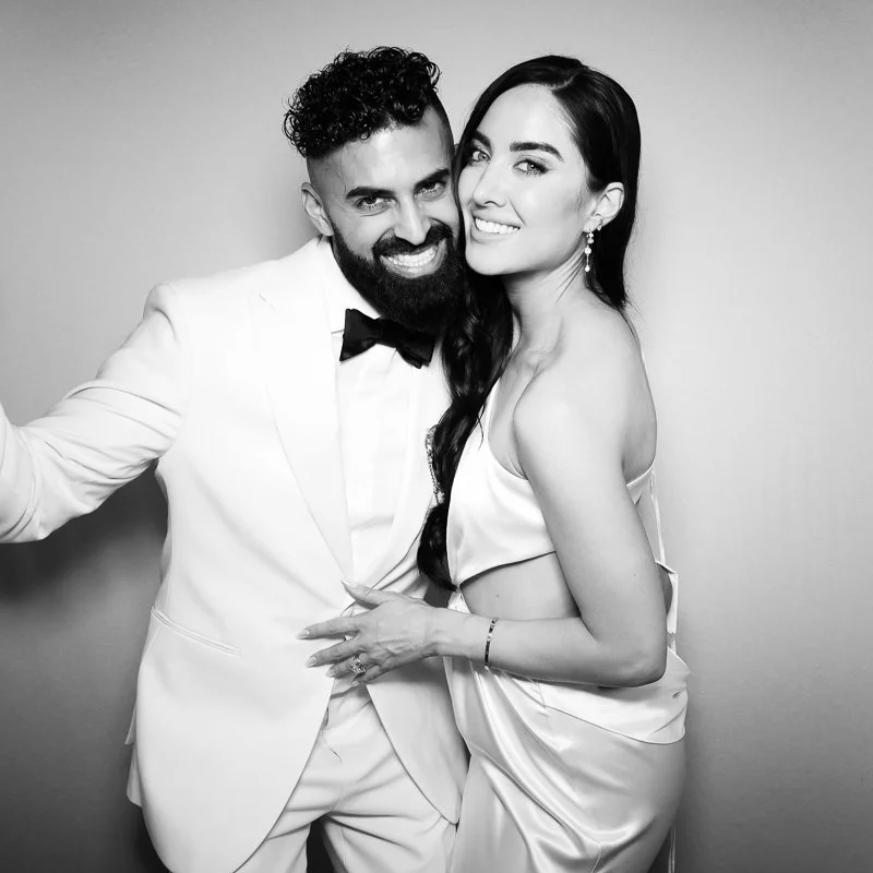 A smiling man in a white tuxedo with a bow tie and a woman in a satin dress, both posing closely together.