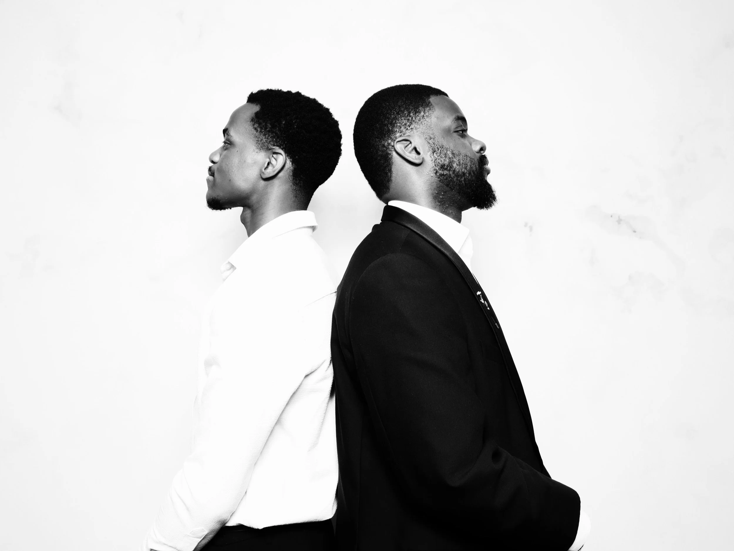 Two men standing back-to-back facing opposite directions, one in a white shirt and the other in a black suit, against a plain background.