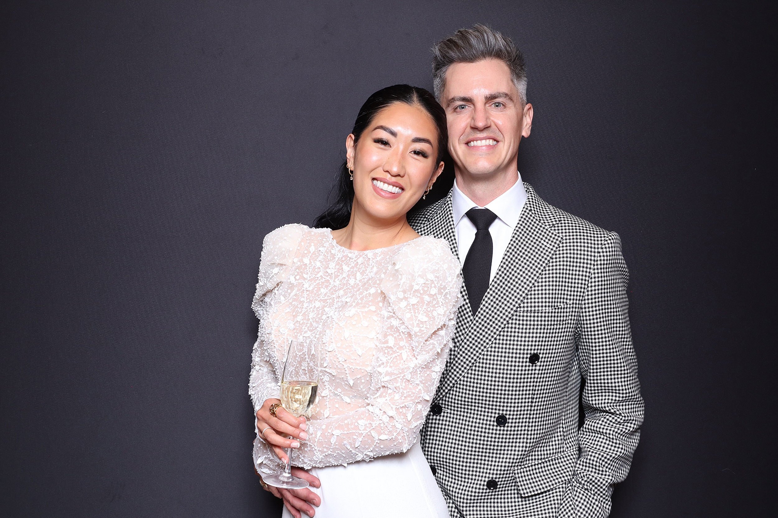 A smiling couple dressed in formal attire, with the woman holding a glass of champagne, standing against a dark background.