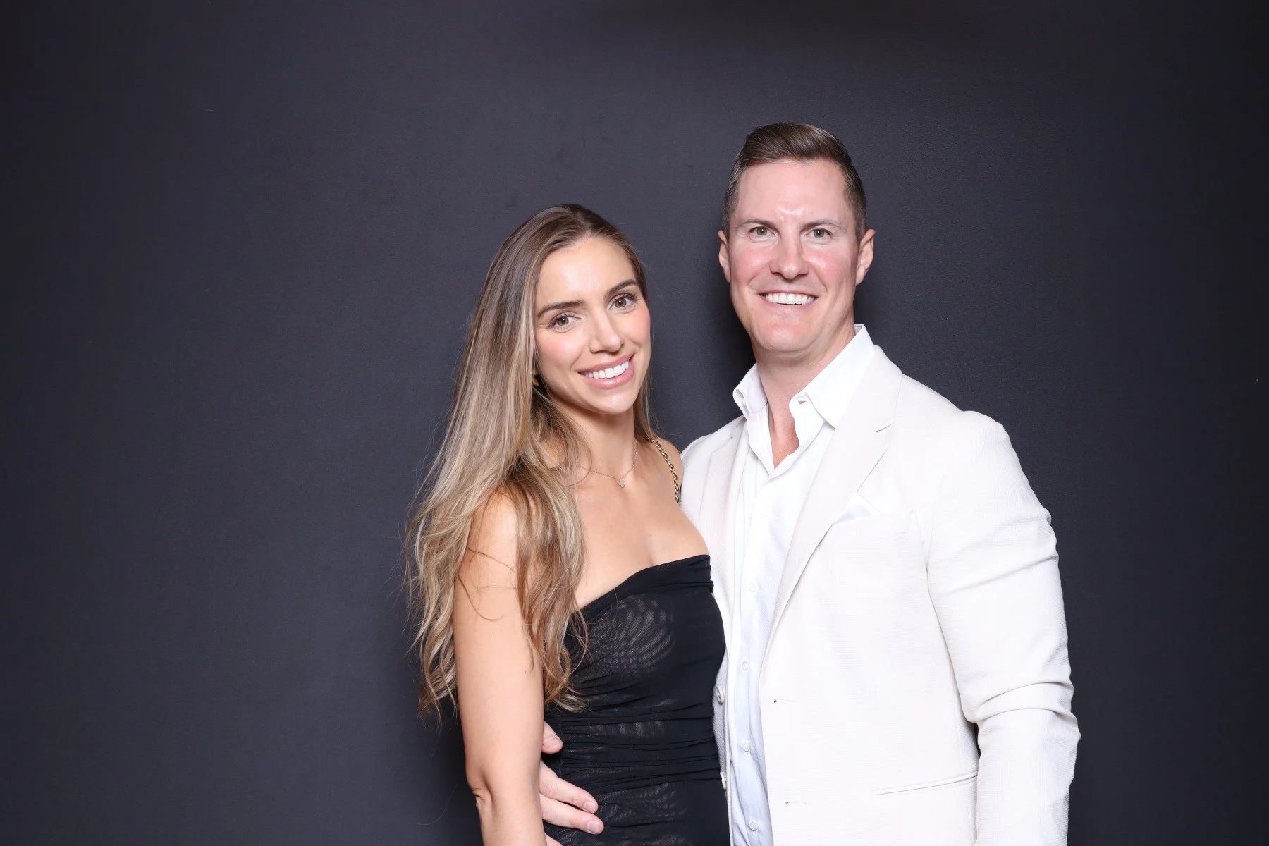 A smiling woman with long wavy hair in a black dress, and a smiling man in a white suit jacket and shirt, standing close against a dark background.