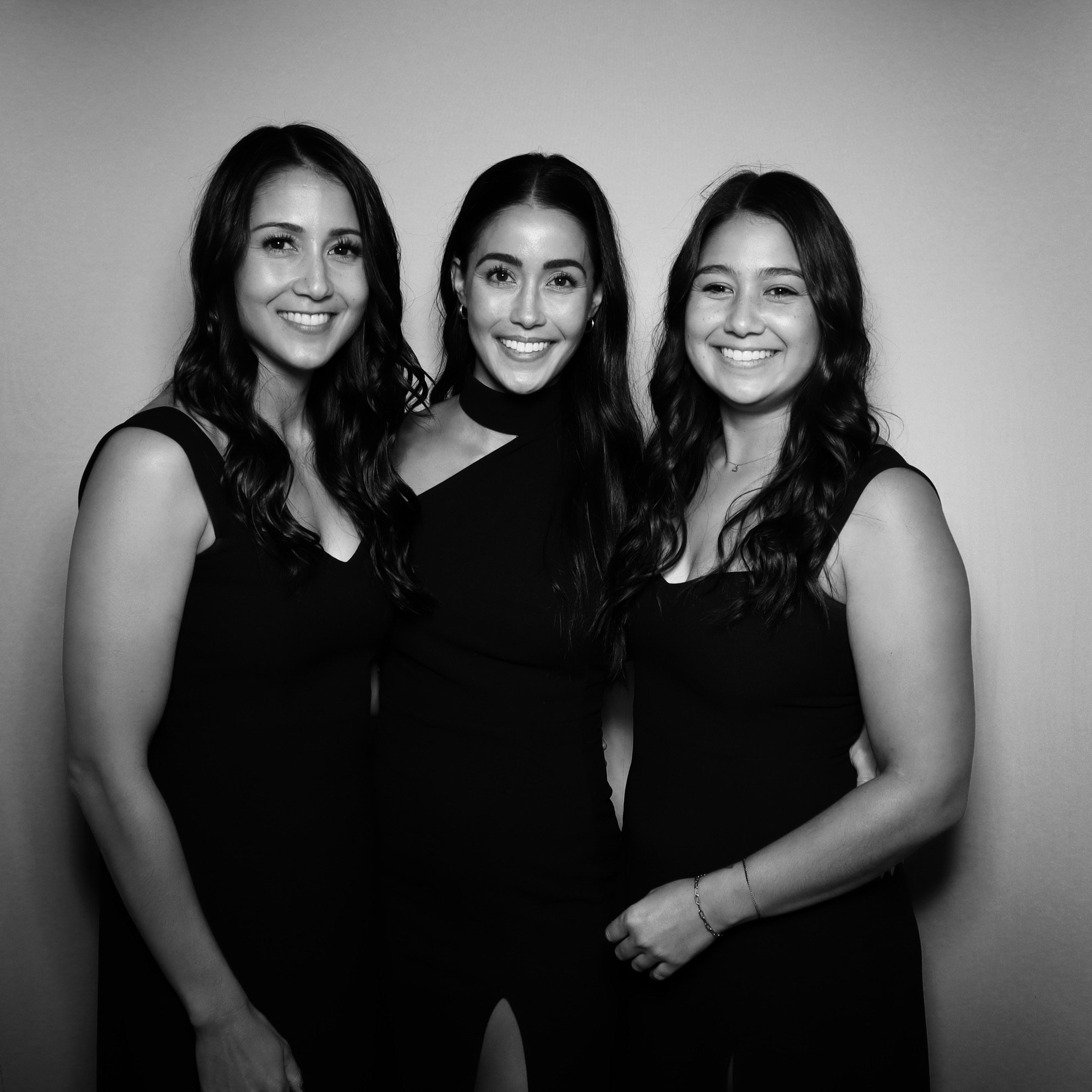 Three young women with long hair smiling, dressed in black, standing close together in front of a plain background.