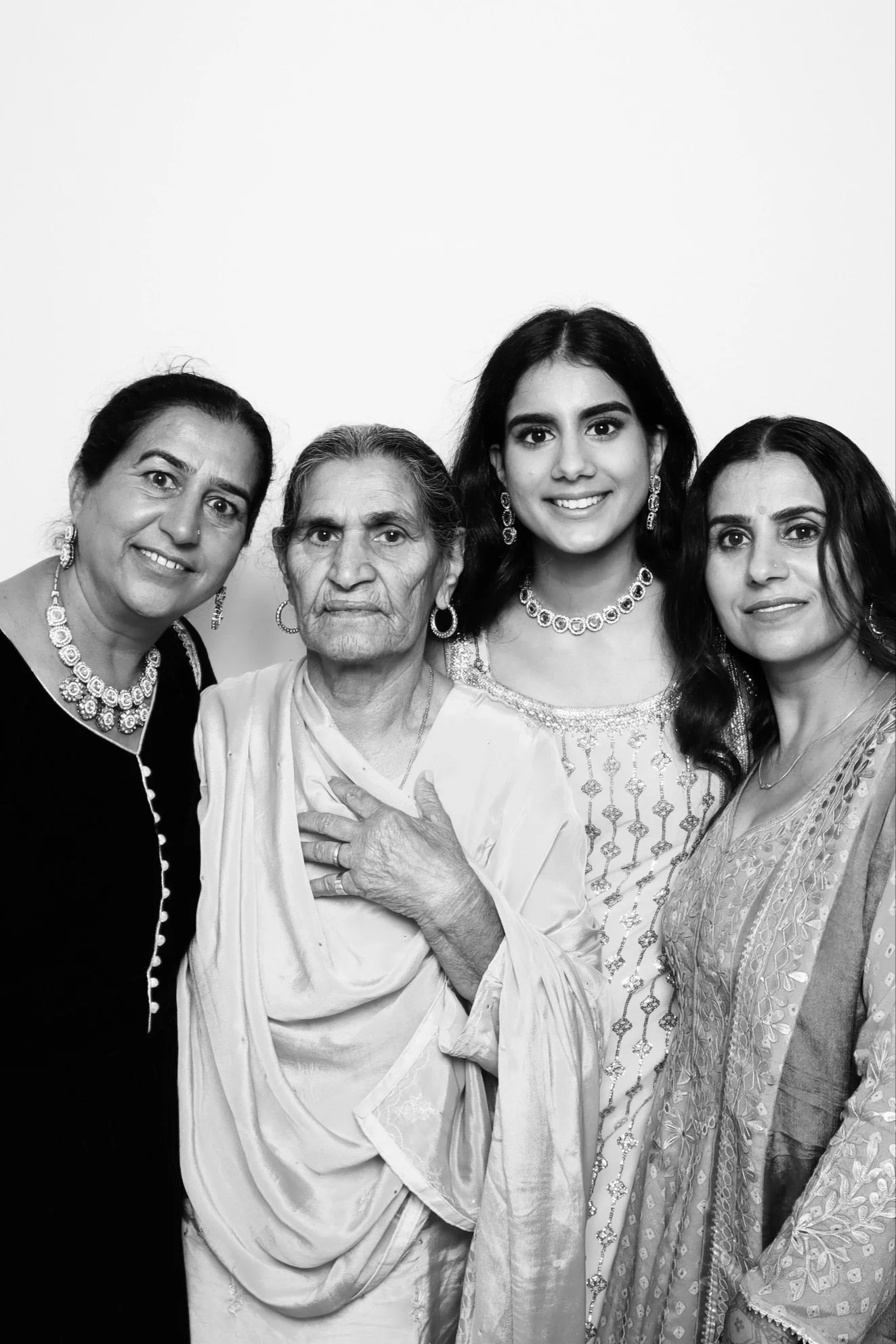 Four women of different ages dressed in traditional attire, wearing jewelry, standing together for a photo against a plain background.