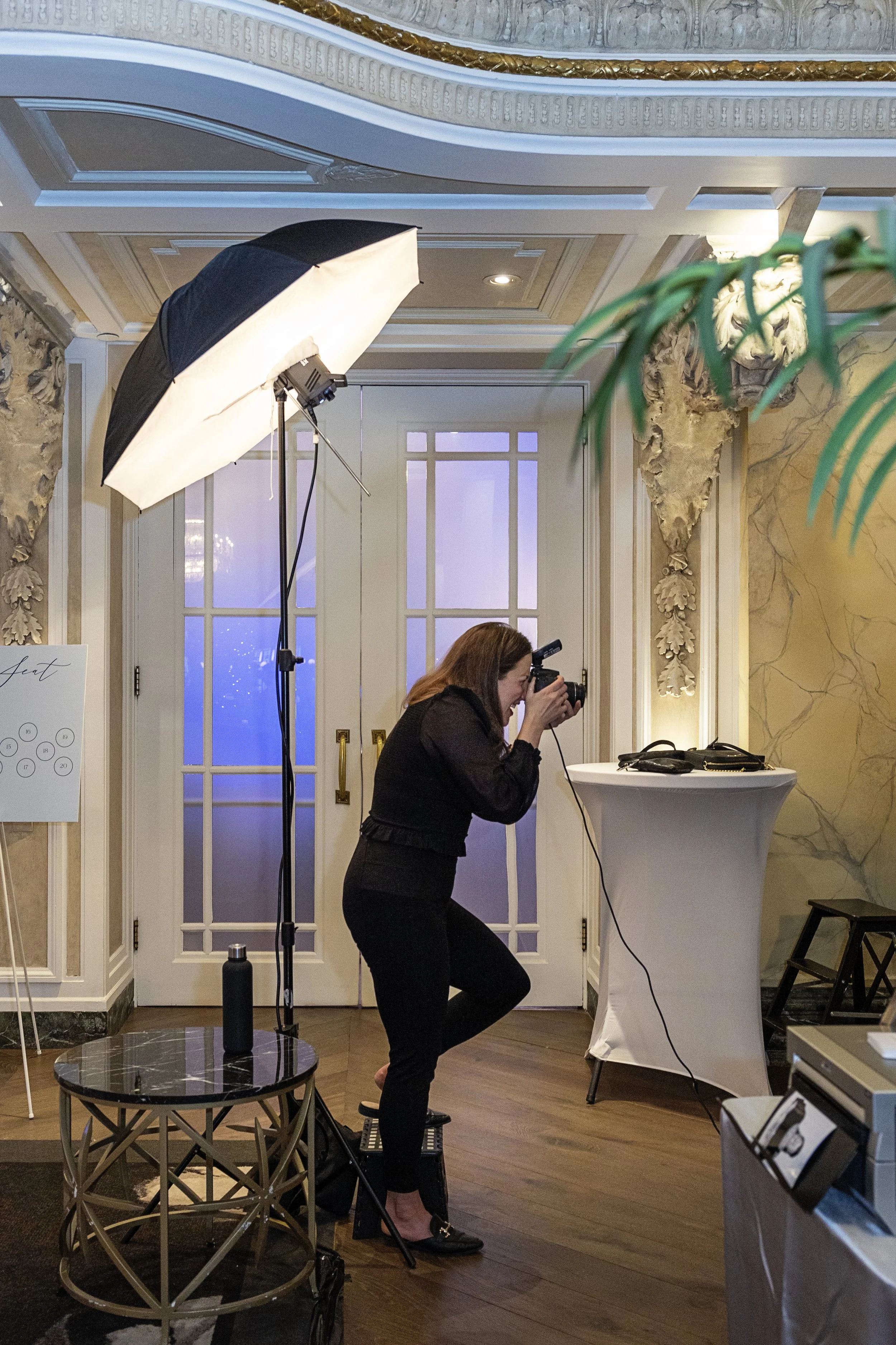A woman taking a photograph indoors using a professional camera. She is standing on a stool in front of a high-top table, with a large umbrella light on her left, and a white double door with glass panels behind her.