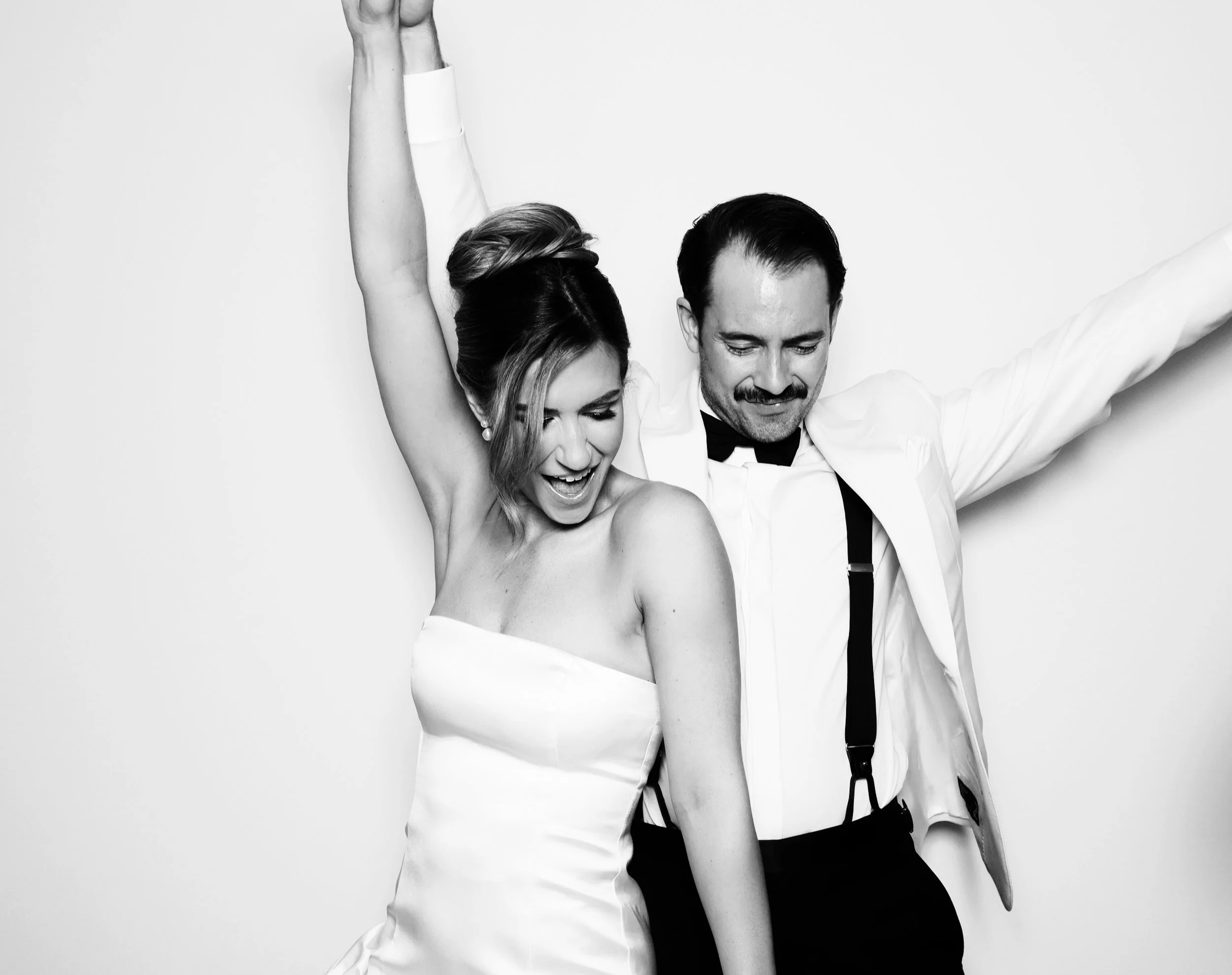 A smiling woman in a strapless dress and a man in a tuxedo with suspenders celebrating together against a plain background.