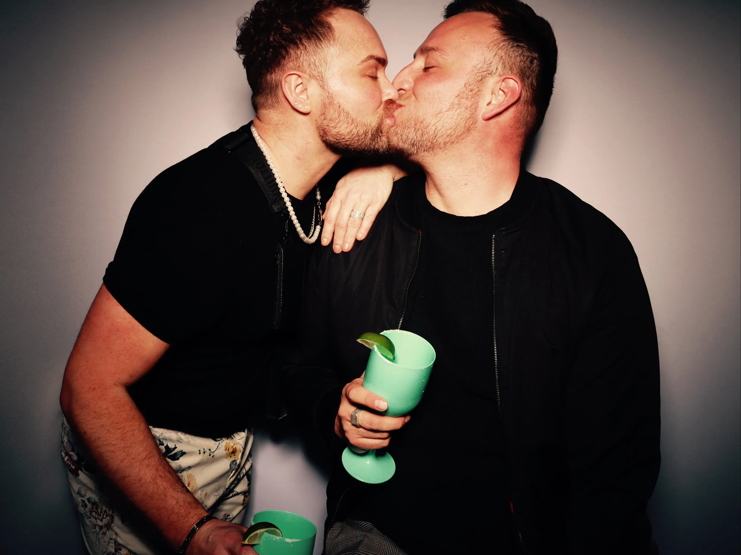 Two men are sharing a kiss while holding green drinks with lime wedges. One man has a pearl necklace and an earring, and the other has a ring on his finger. They are dressed in black and are against a plain background.