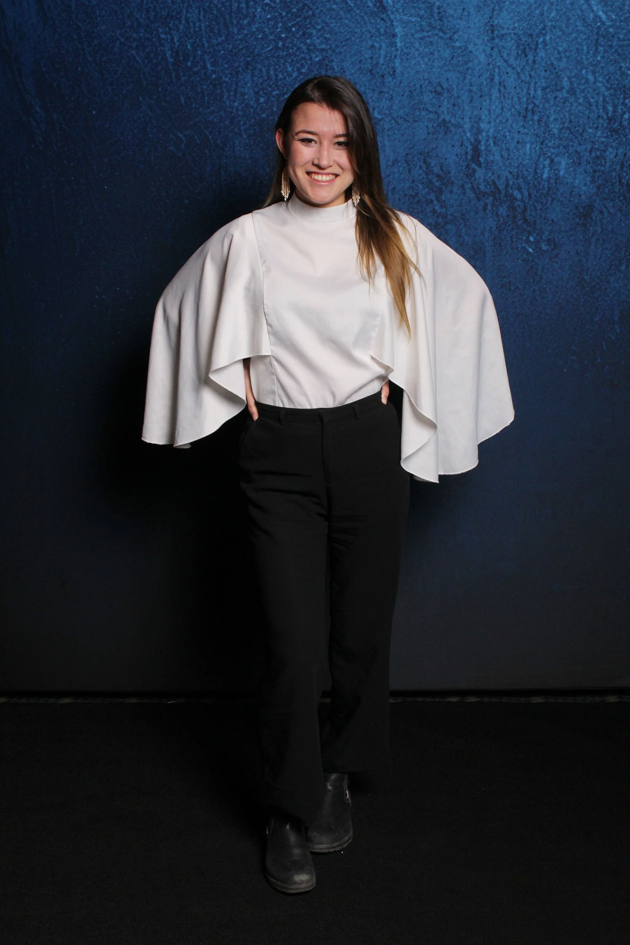 Young woman with long hair and earrings, wearing a white top with cape-like sleeves, black pants, and black boots, standing against a blue textured wall and smiling.