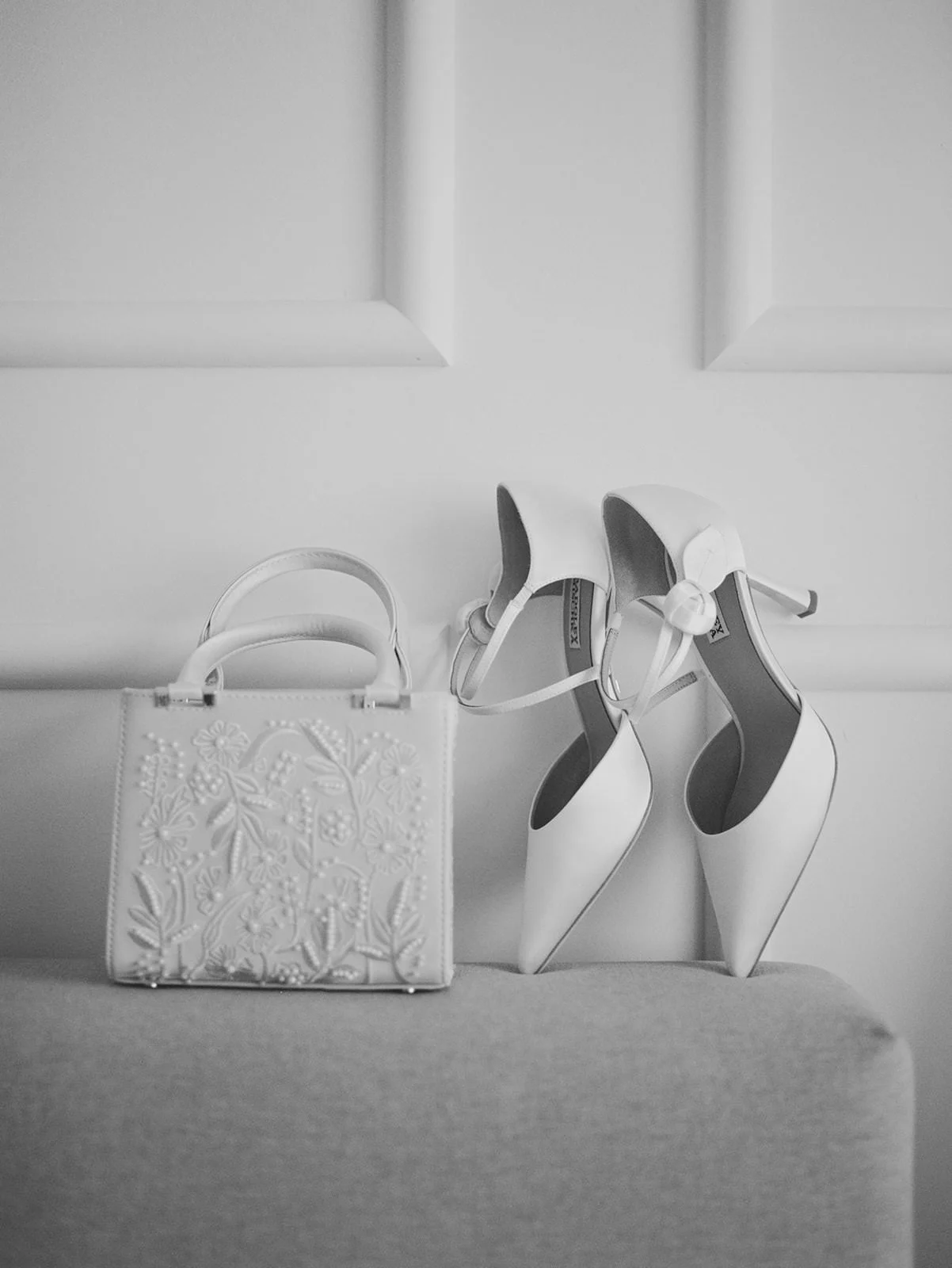 A pair of white high-heeled shoes with ankle straps and a white handbag with floral embroidery, both resting on a sofa.