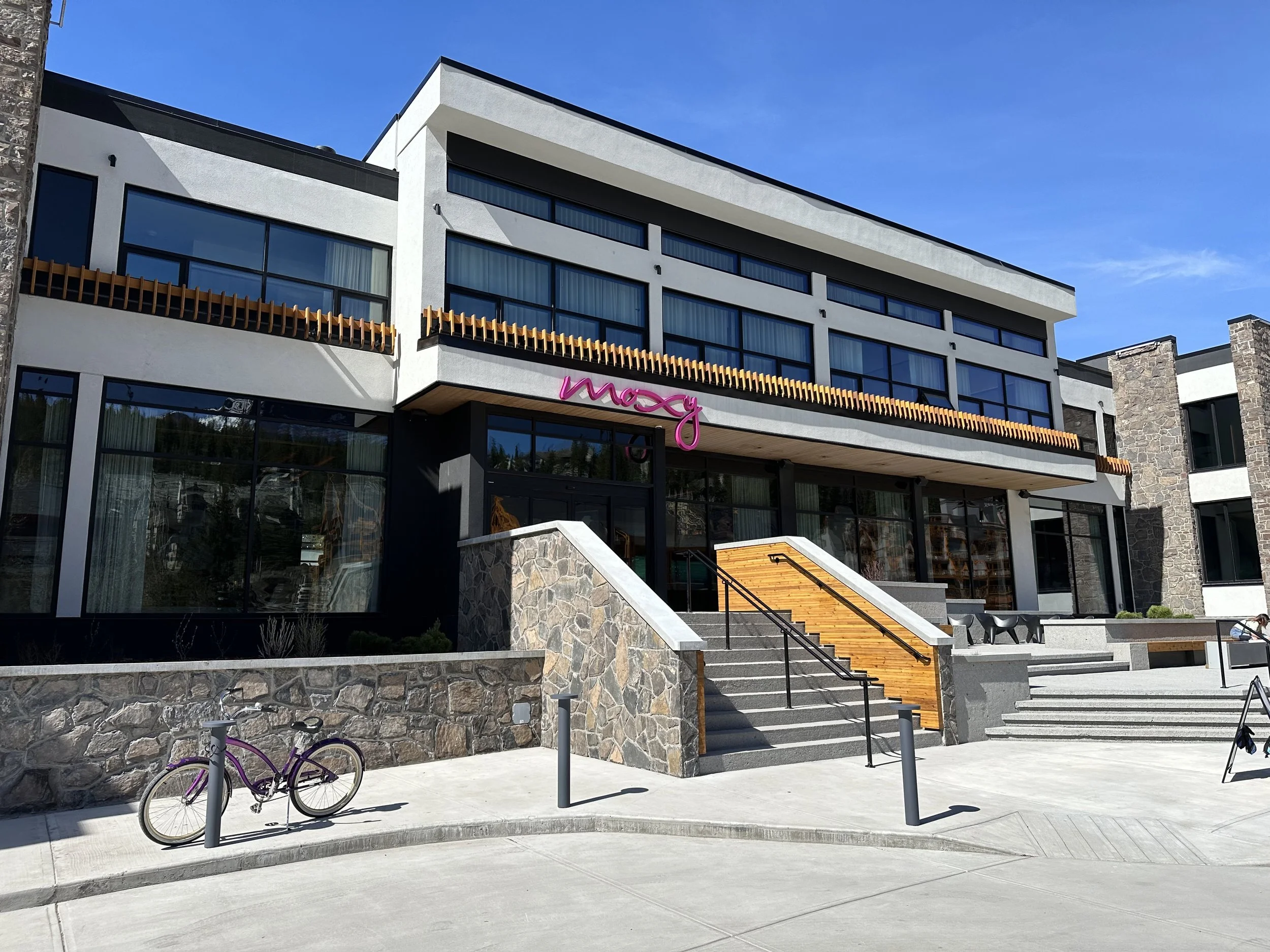 Modern building with large glass windows, stone accents, wooden railings, and a purple bicycle in front. The sign says 'macy' in pink cursive letters.