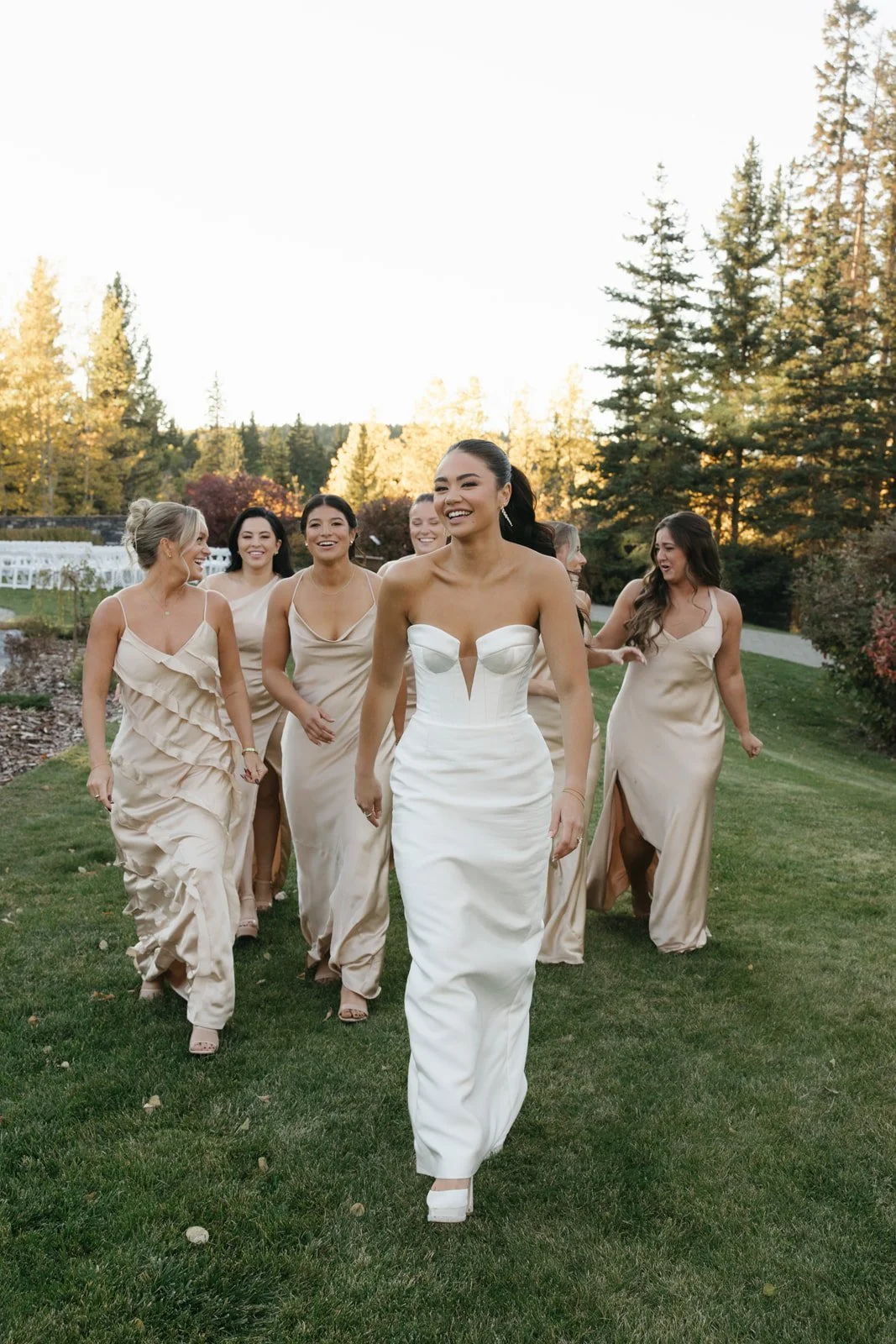 Group of women in elegant dresses walking outdoors, smiling and laughing, with trees and a clear sky in the background.