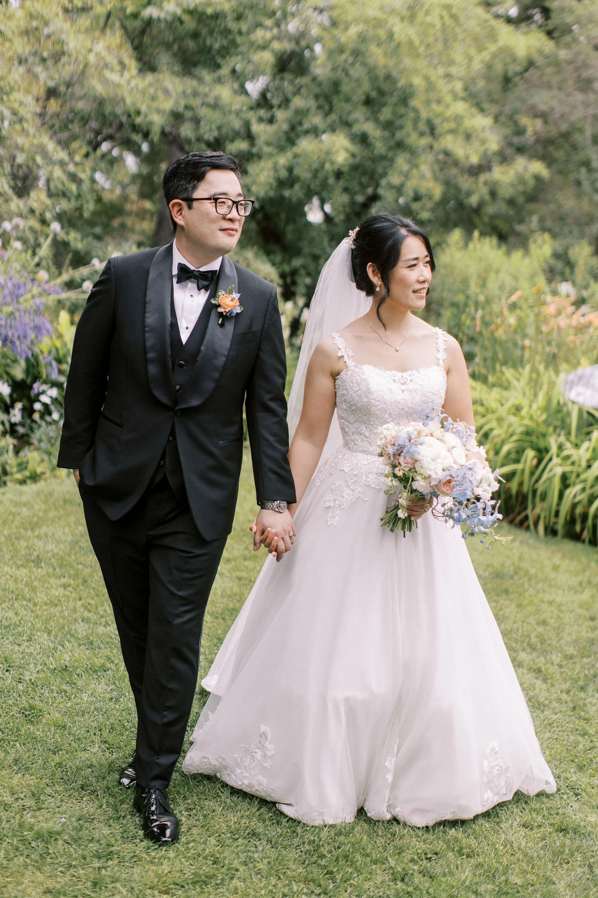 A bride and groom walking outdoors on a wedding day, holding hands, with greenery and flowers in the background.