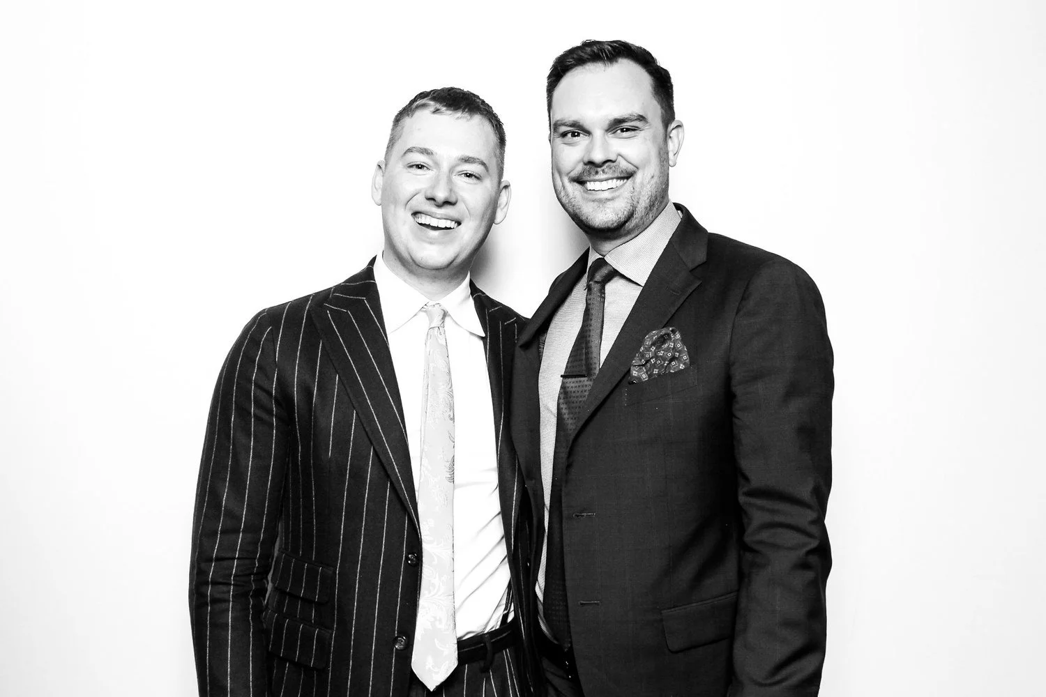 Two smiling men in suits standing close together, posing for a photo, on a plain background.