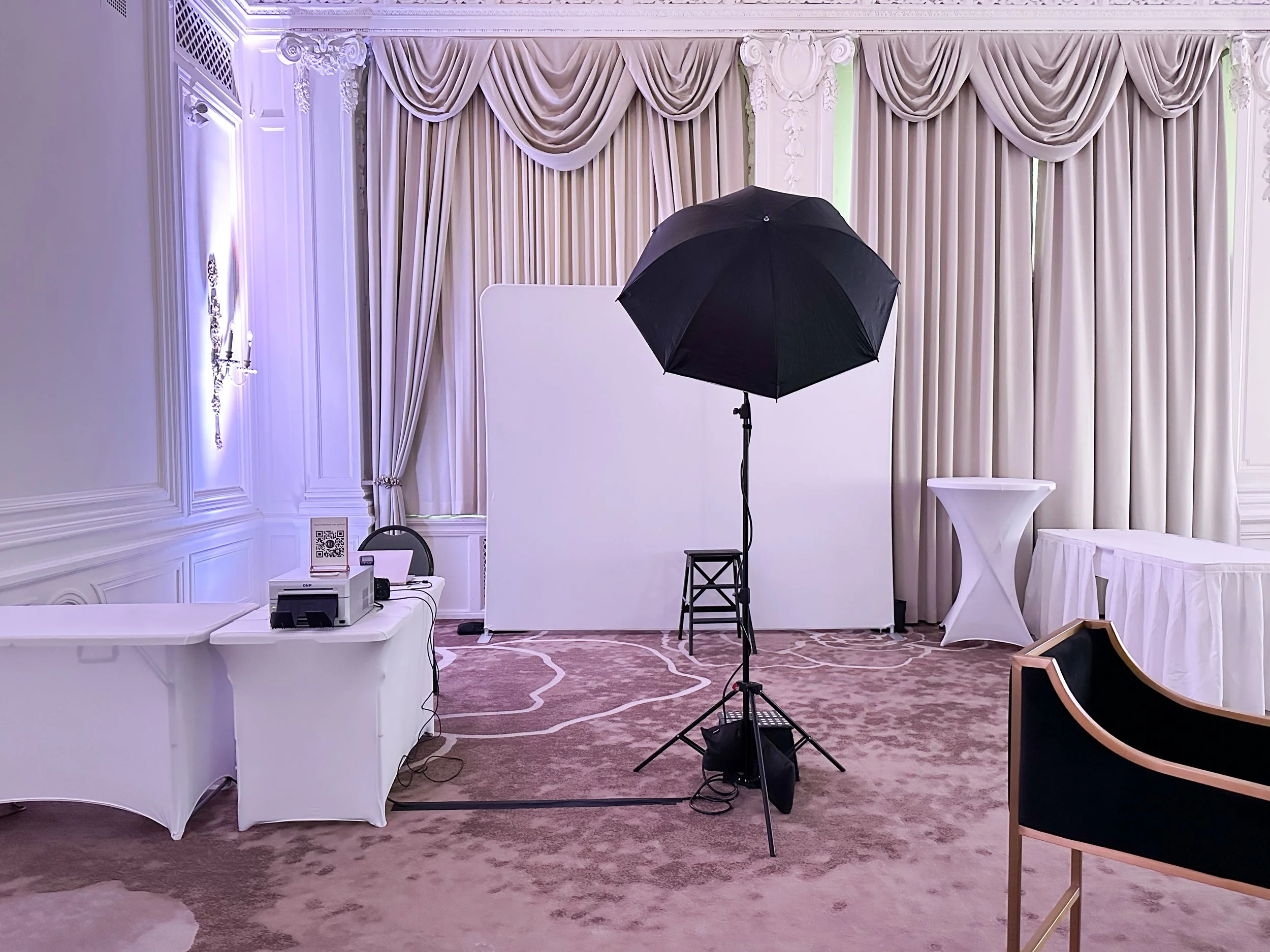Photo of a professional photography setup in an elegant room with white walls and curtains, featuring a large umbrella light, a white backdrop, a small table with camera equipment, and chairs.