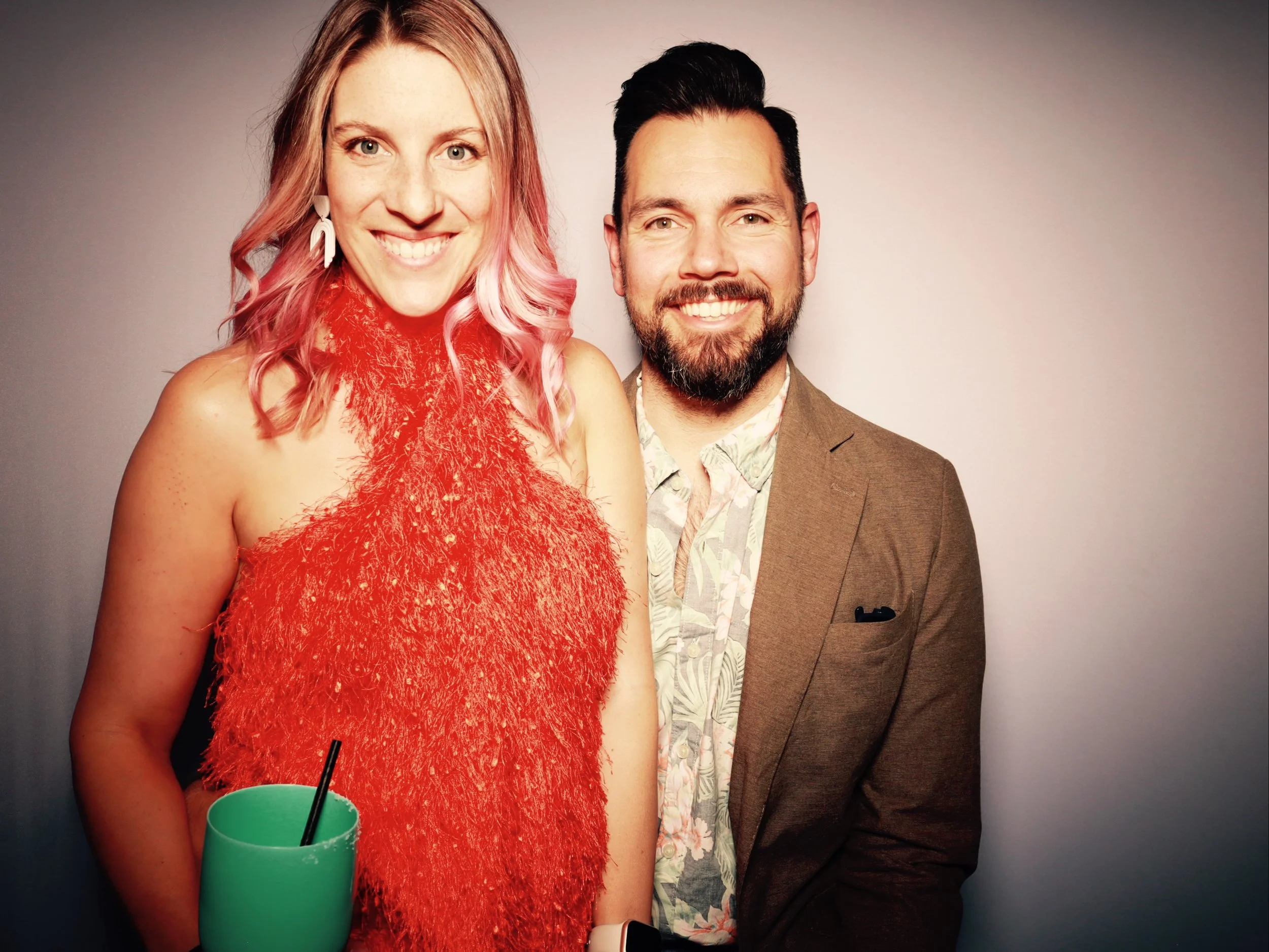 A smiling woman with pinkish-blonde hair wearing a red, fuzzy sleeveless top holding a green cup with a straw, standing next to a smiling man with dark hair and a beard wearing a brown blazer and a floral shirt, against a plain background.