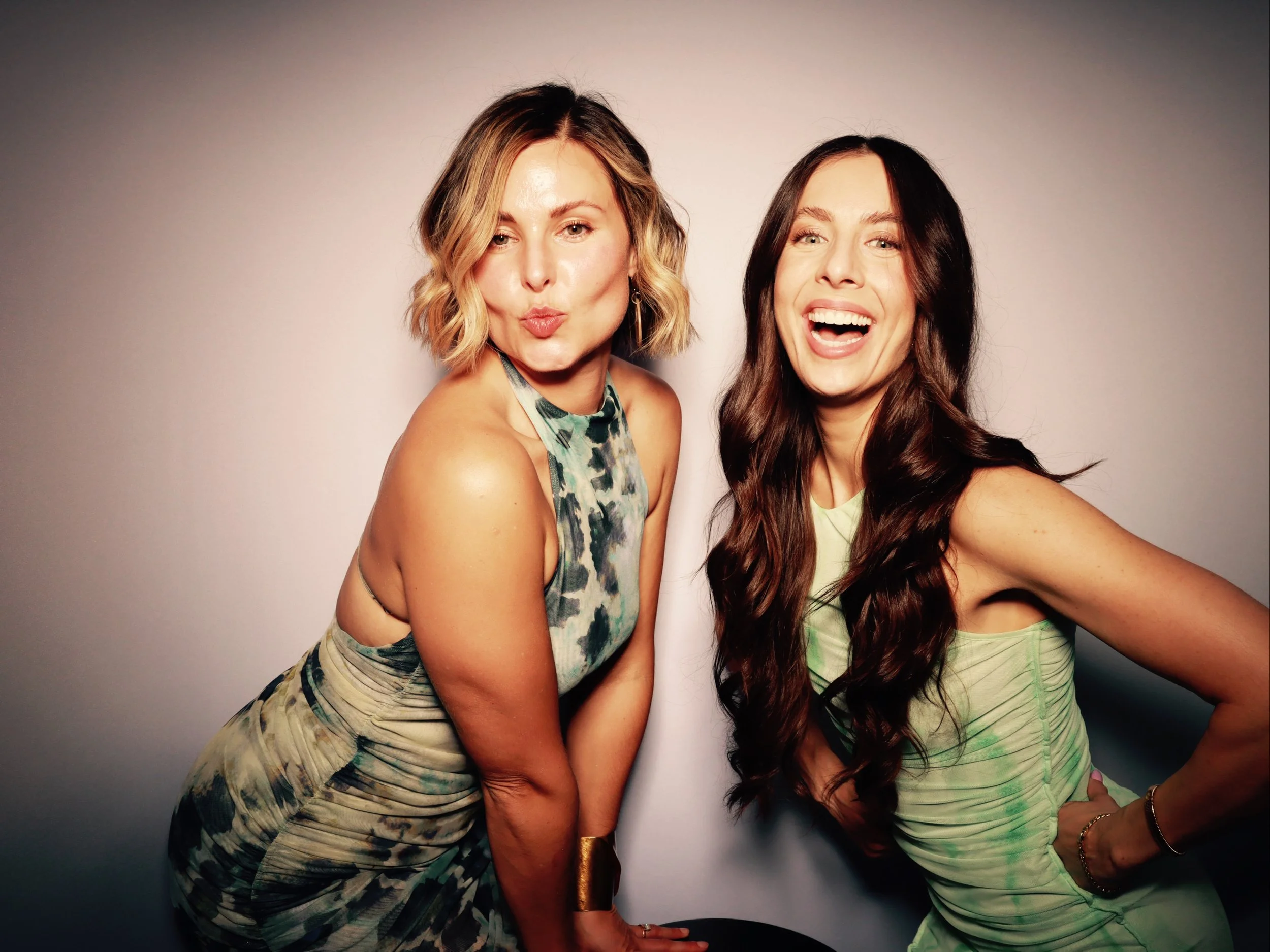 Two women posing for a photo, one with short wavy blonde hair in a tie-dye dress making a kissy face and the other with long dark hair in a light green dress smiling widely with her arm on her hip, against a gradient background.