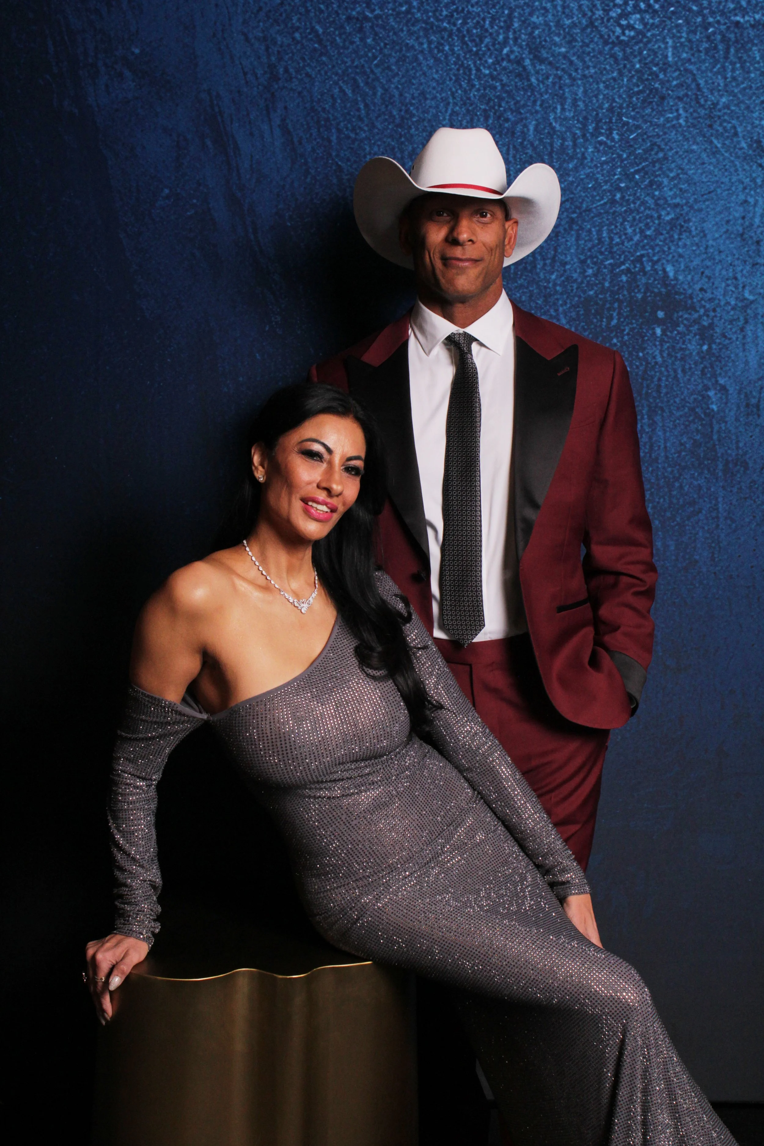A woman in a shimmering, off-the-shoulder gown and a man in a maroon suit with black lapels, white shirt, patterned tie, and white cowboy hat, posing against a textured blue background.