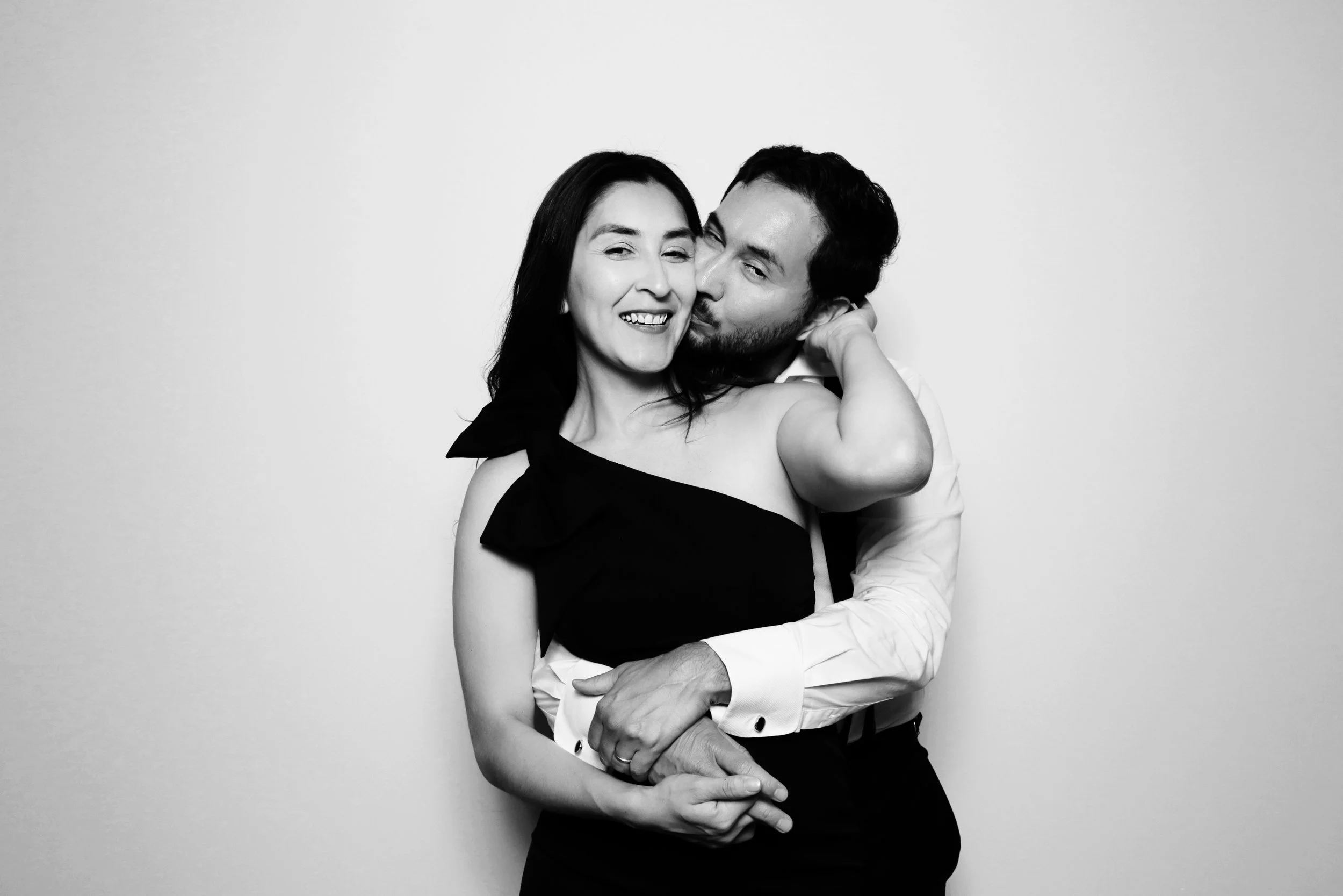 A black and white photo of a happy couple, with the woman smiling and the man kissing her cheek, against a plain background.