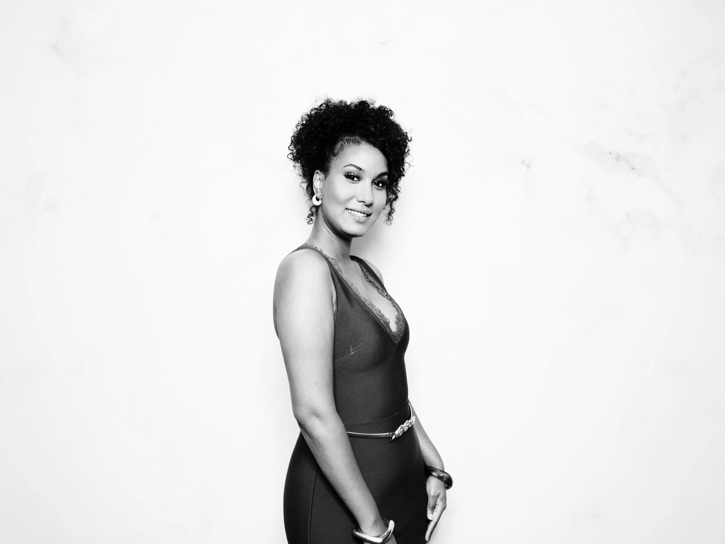 Black and white portrait of a woman with curly hair, wearing a dress with lace details, earrings, and bracelets, standing against a plain background.