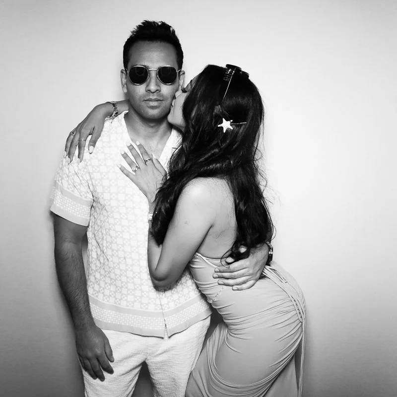 A man and woman pose together, with the woman kissing the man's cheek. The man is wearing sunglasses and a patterned short-sleeve shirt, while the woman has long dark hair, a headband, and a fitted dress.