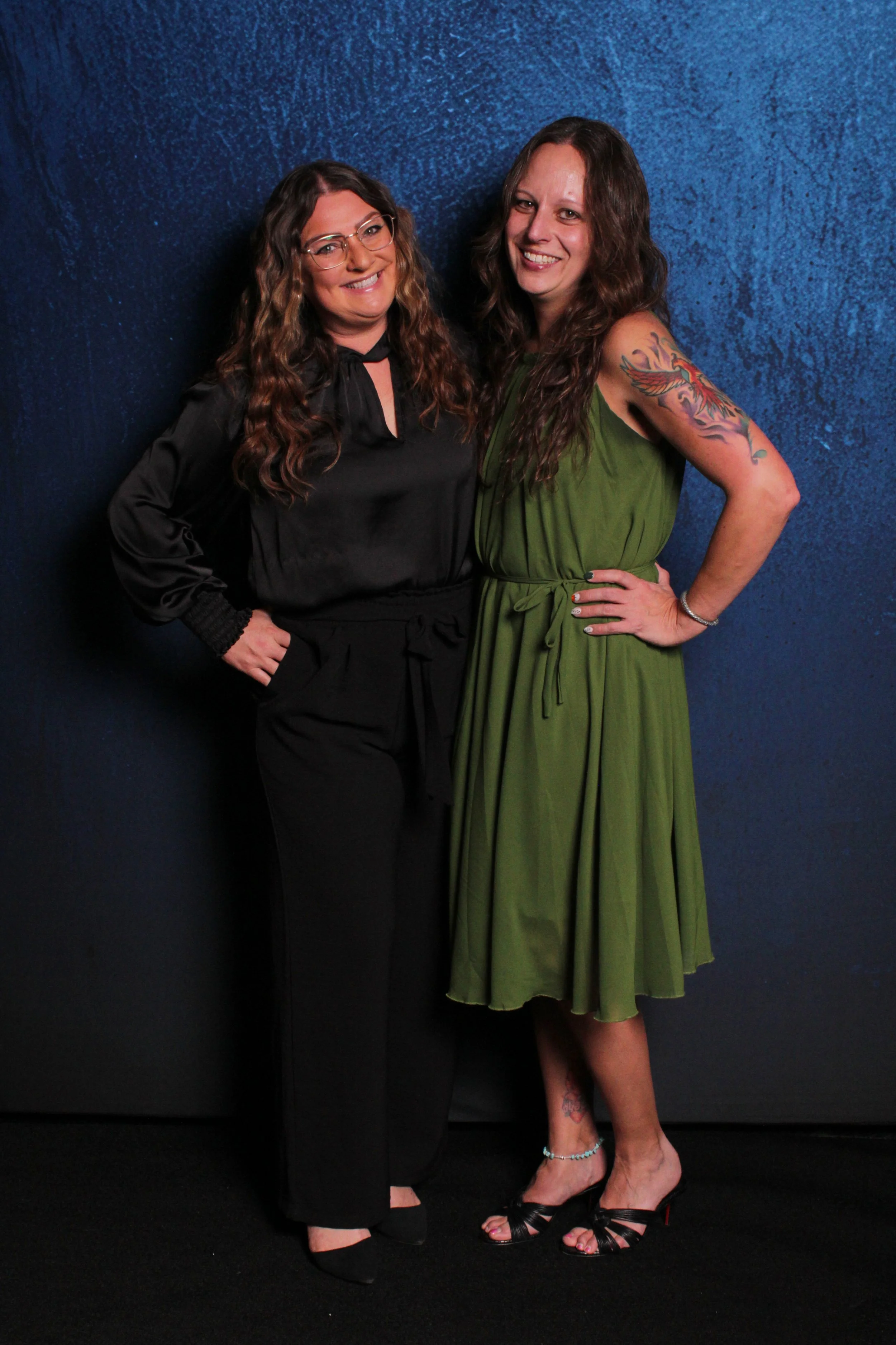 Two women standing together in front of a blue textured wall, smiling at the camera. The woman on the left is wearing black glasses and a black outfit with long wavy brown hair. The woman on the right is wearing a green dress with a tattoo on her upp