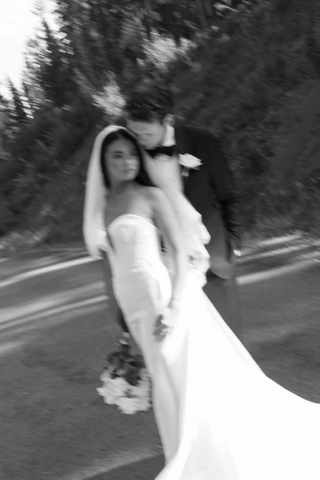 Blurry black and white photo of a bride in a strapless wedding gown holding a bouquet, and a groom in a tuxedo with a bow tie and boutonniere, walking together outdoors.