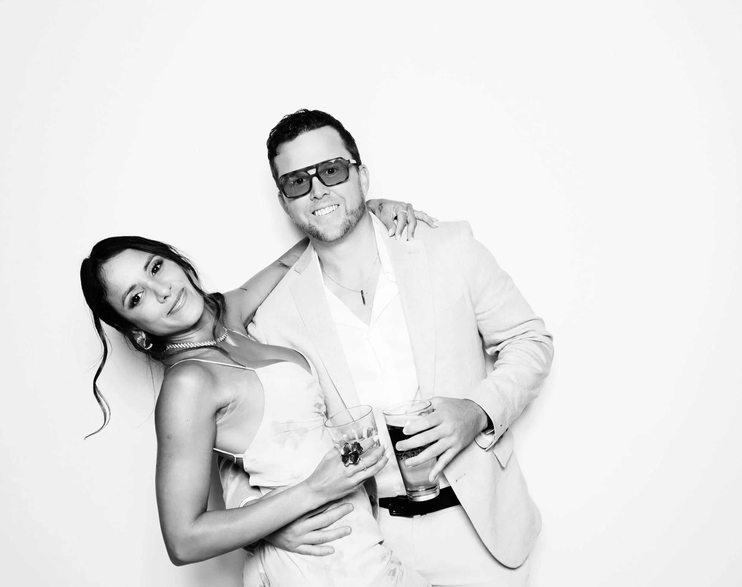 A smiling couple holding drinks at a party. The woman has dark hair, wearing a dress, necklace, and earrings. The man wears glasses, a blazer, and a watch. They stand against a plain white background.