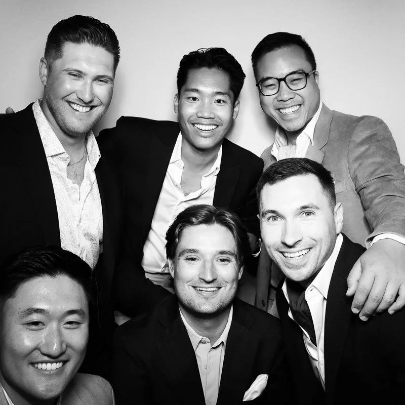 Group of six men dressed in business casual attire smiling and posing together against a plain background.