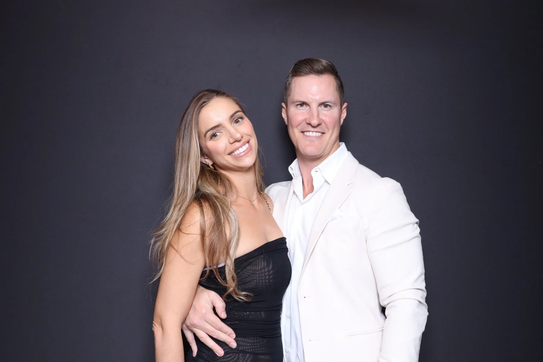 A smiling couple, the woman with long light brown hair in a black strapless dress and the man with short dark hair in a white suit, standing close together against a dark background.