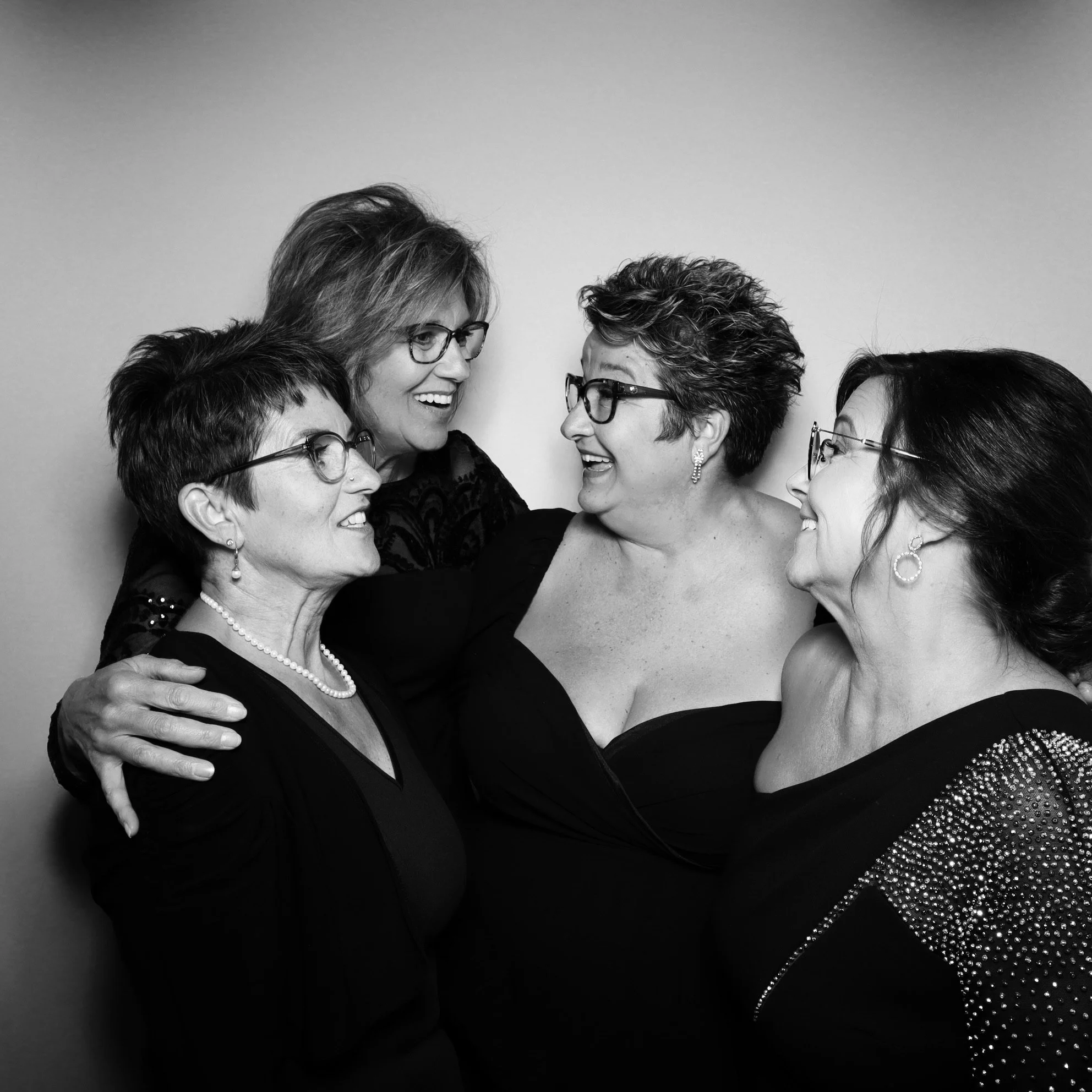 Four women smiling and embracing each other, wearing black dresses and glasses in a close-up portrait.