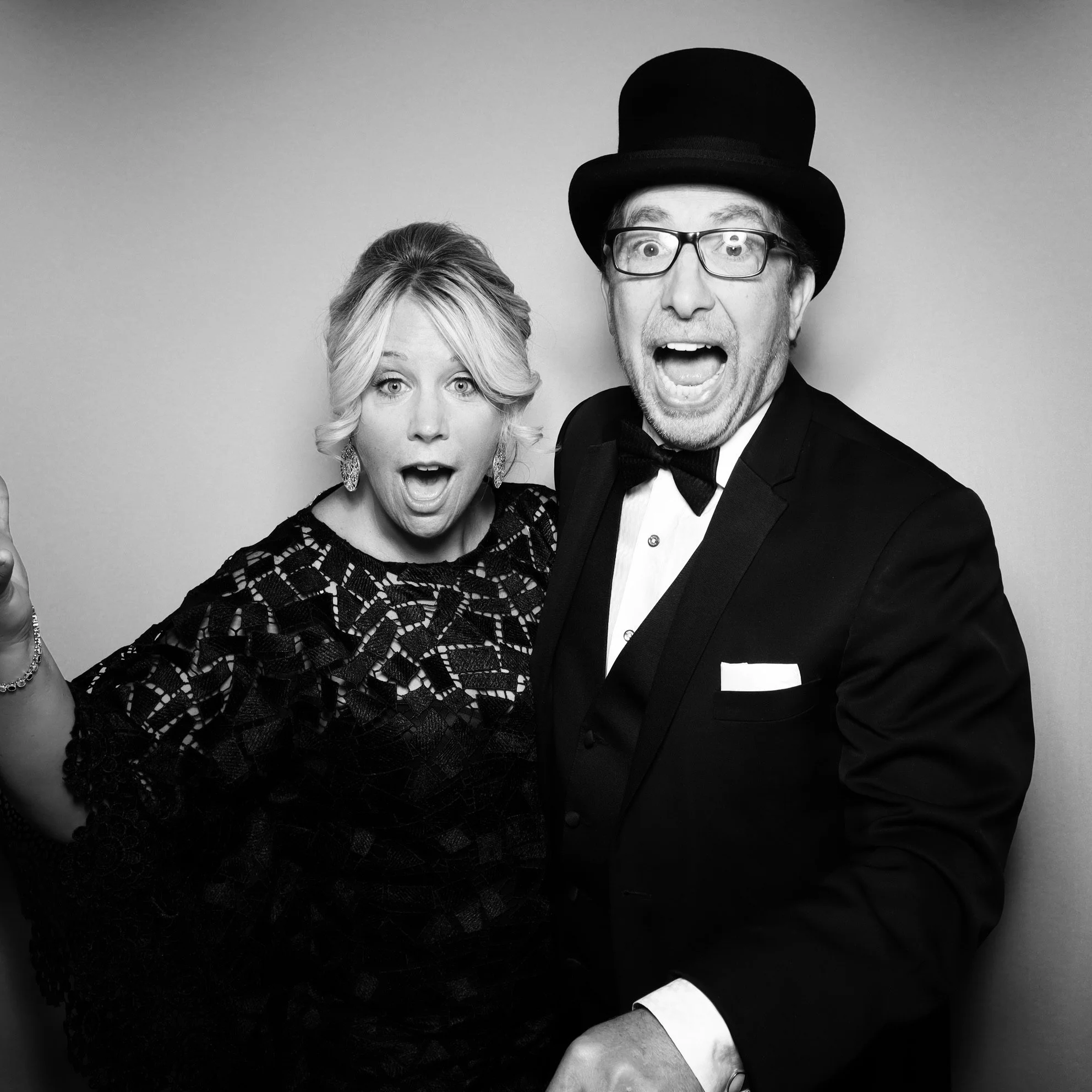 Black and white photo of a surprised woman and a man wearing glasses, a top hat, and a tuxedo, both showing expressions of excitement.